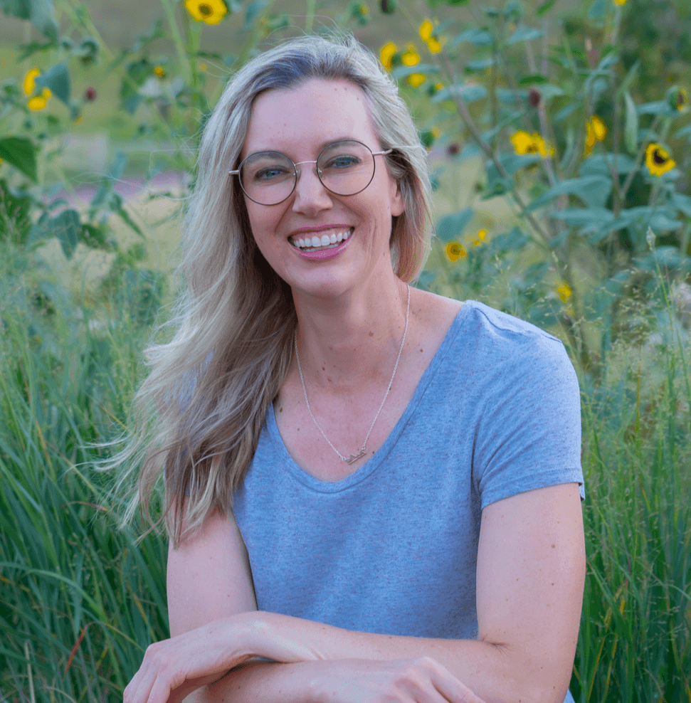 Woman with glasses and long blonde hair smiling outdoors among sunflowers and greenery.