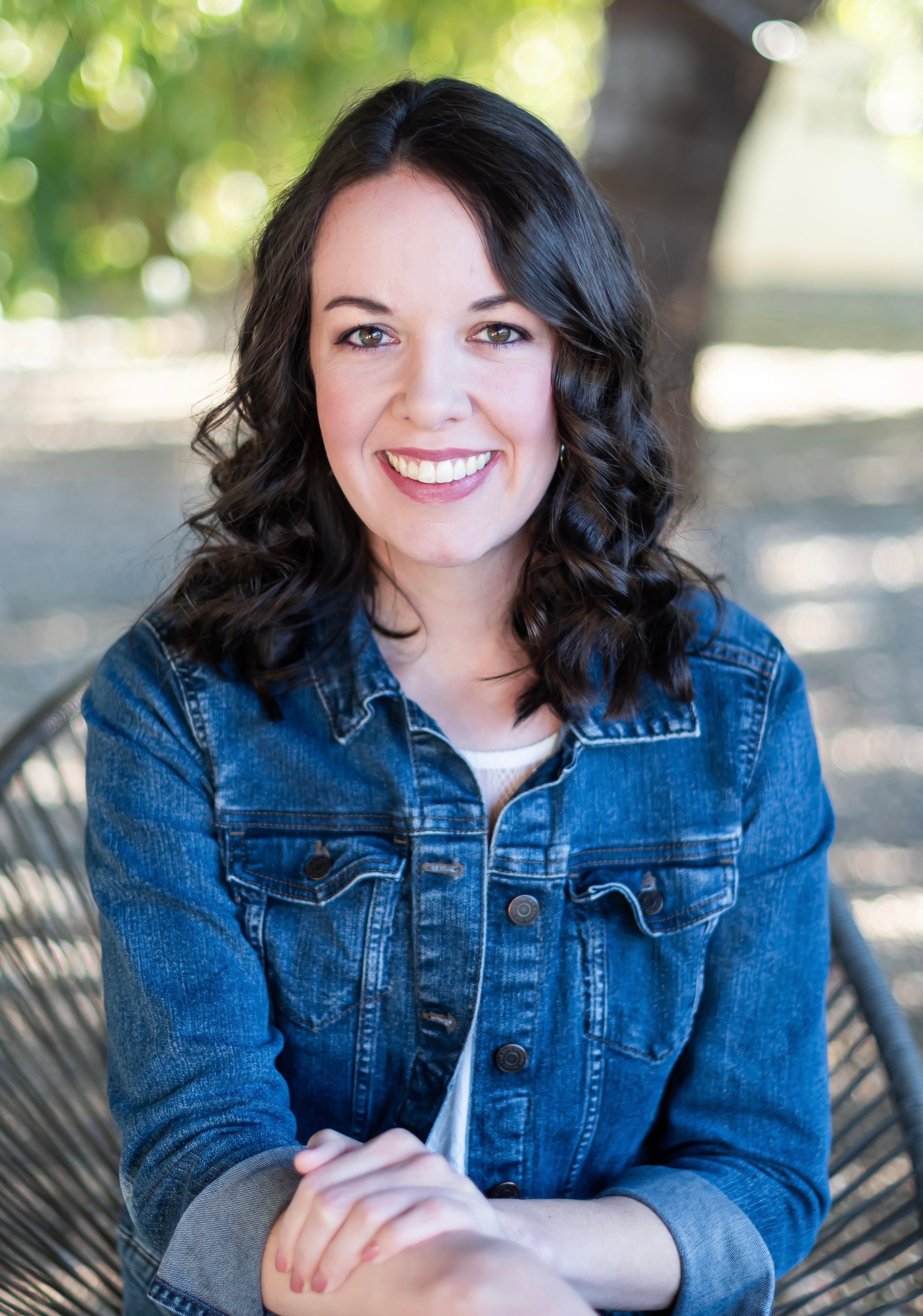 Young woman with dark wavy hair smiling outdoors, wearing a denim jacket, arms crossed, trees in background.