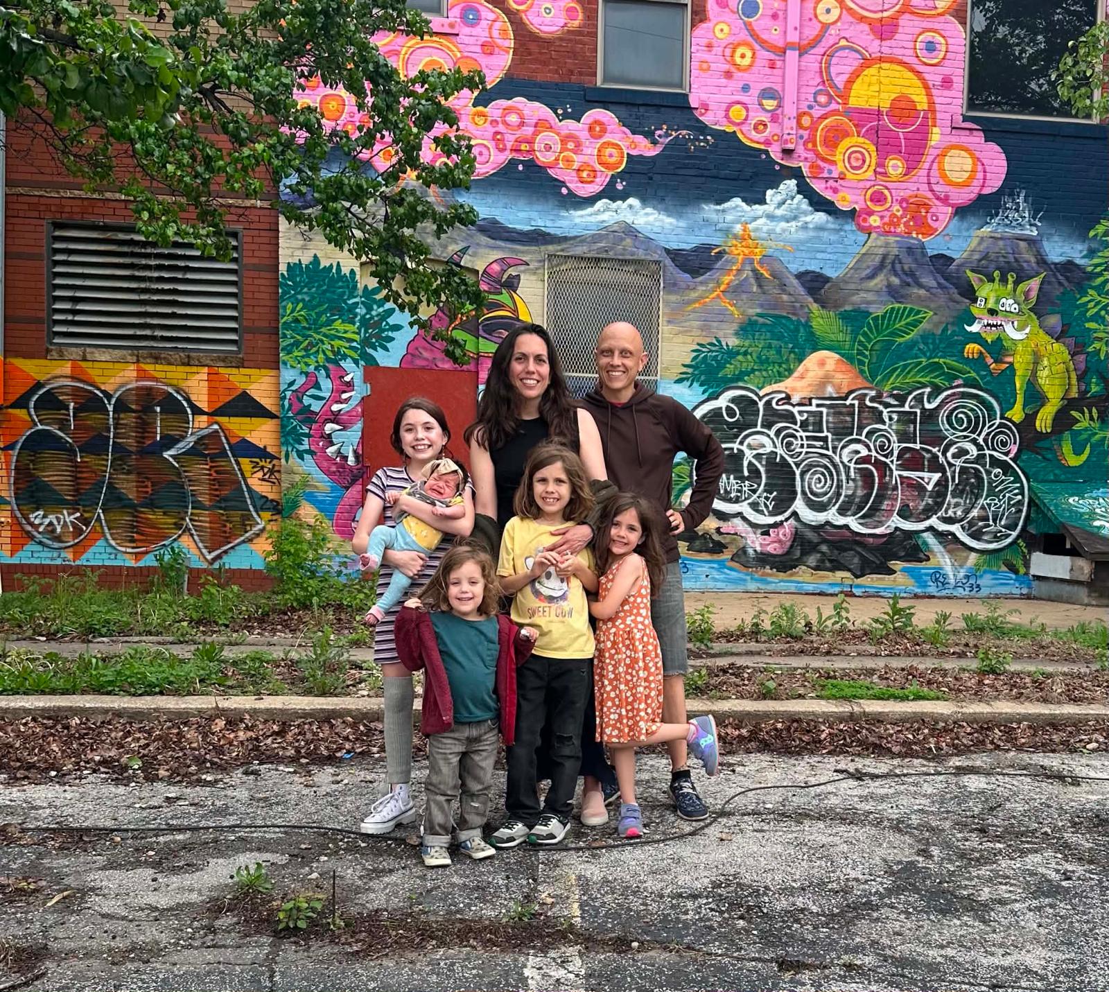 Group of six people, including children and adults, standing in front of a colorful mural with trees and graffiti.