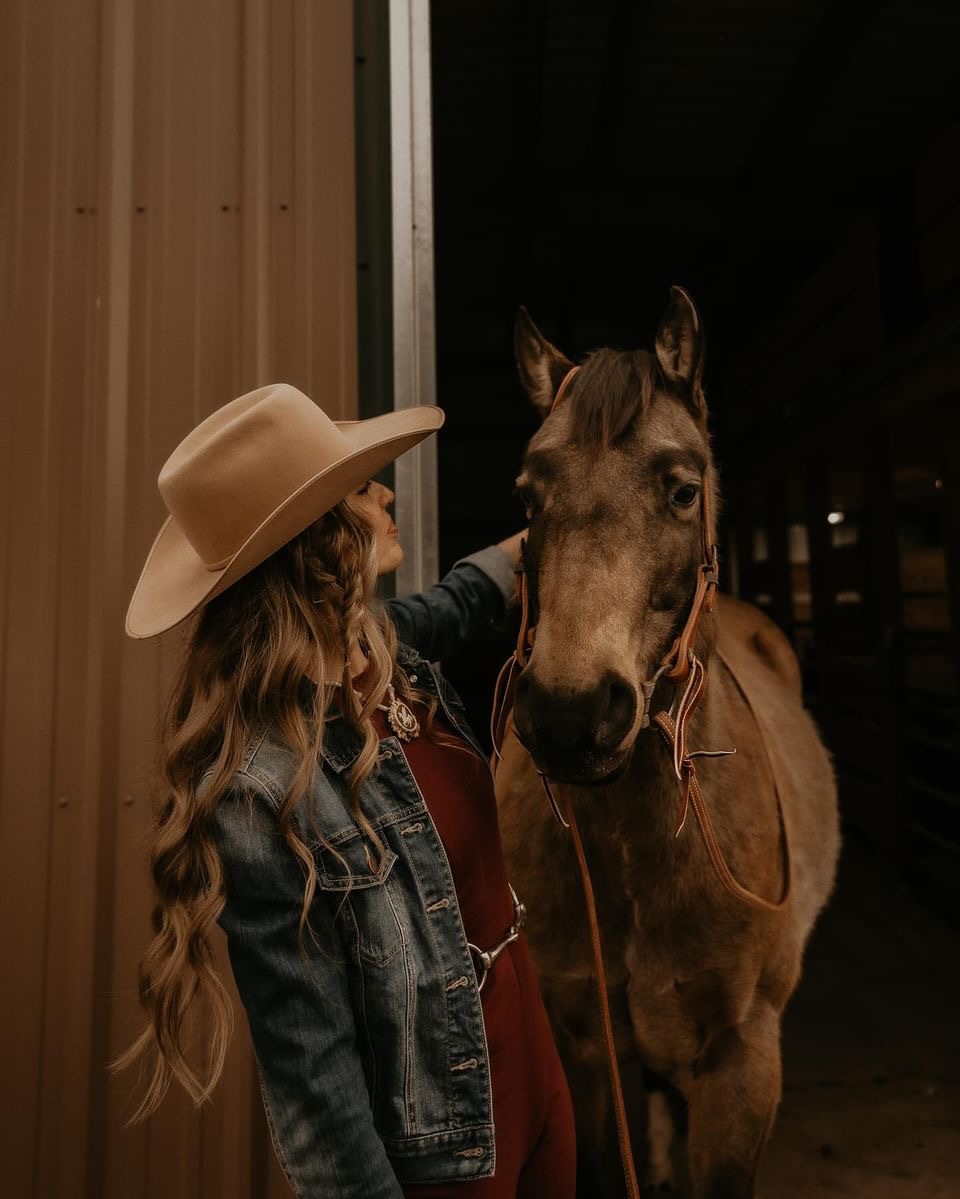 Person with long hair and wide-brimmed hat standing next to a horse inside a barn at night.