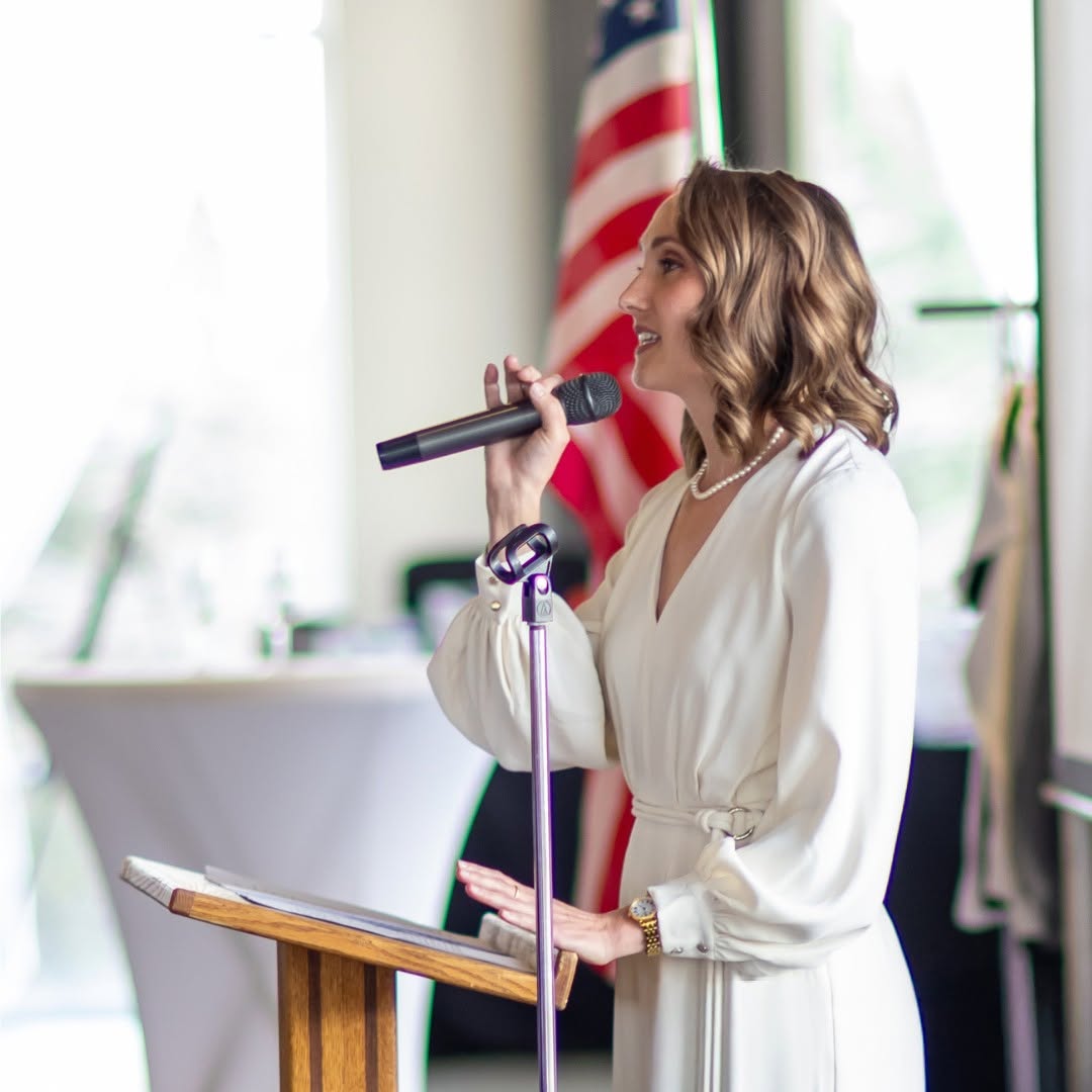 Woman speaking into microphone at podium with American flag in background, wearing white dress, smiling, in a bright room.
