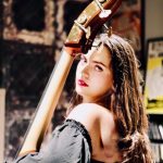 Young woman with long hair holding a cello, looking at the camera, in a dimly lit room with posters and shelves.