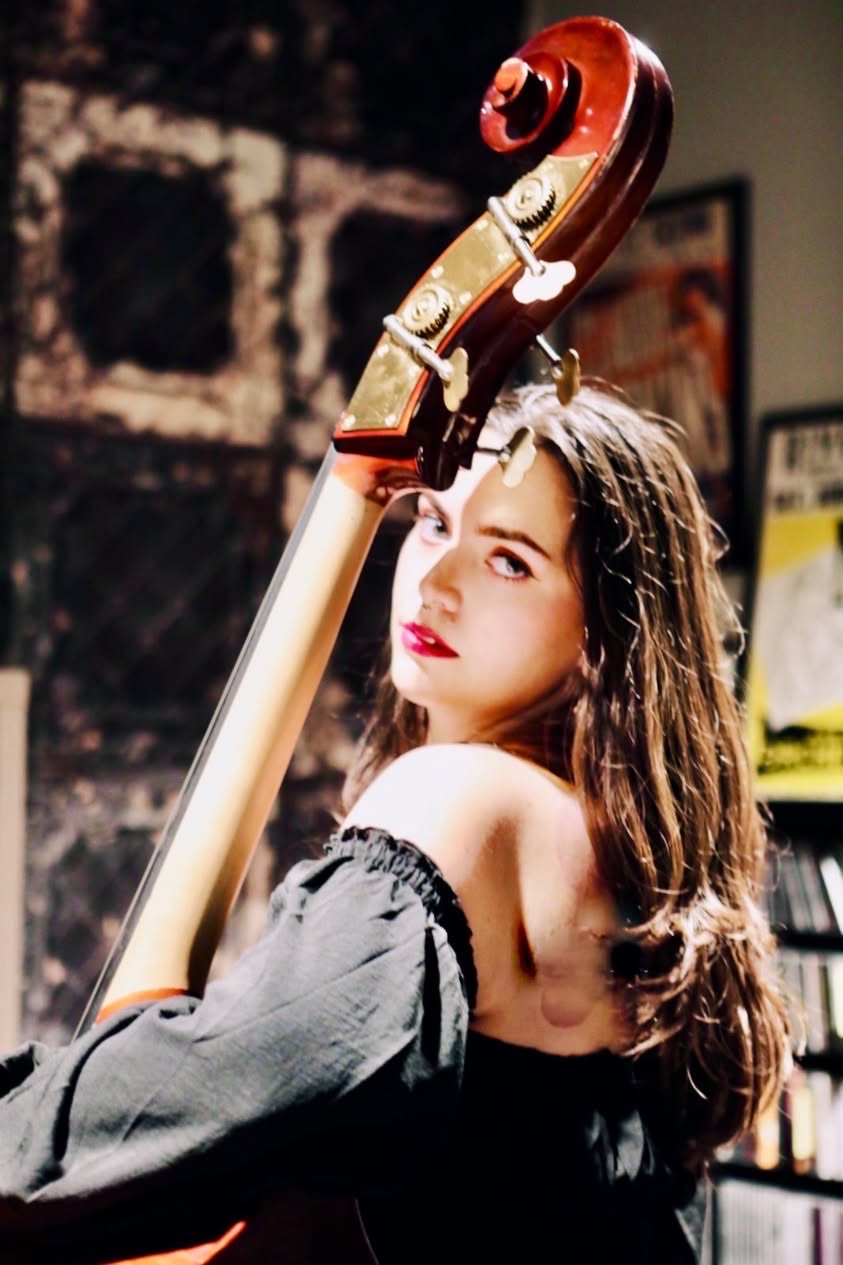 Young woman with long hair holding a cello, looking at the camera, in a dimly lit room with posters and shelves.