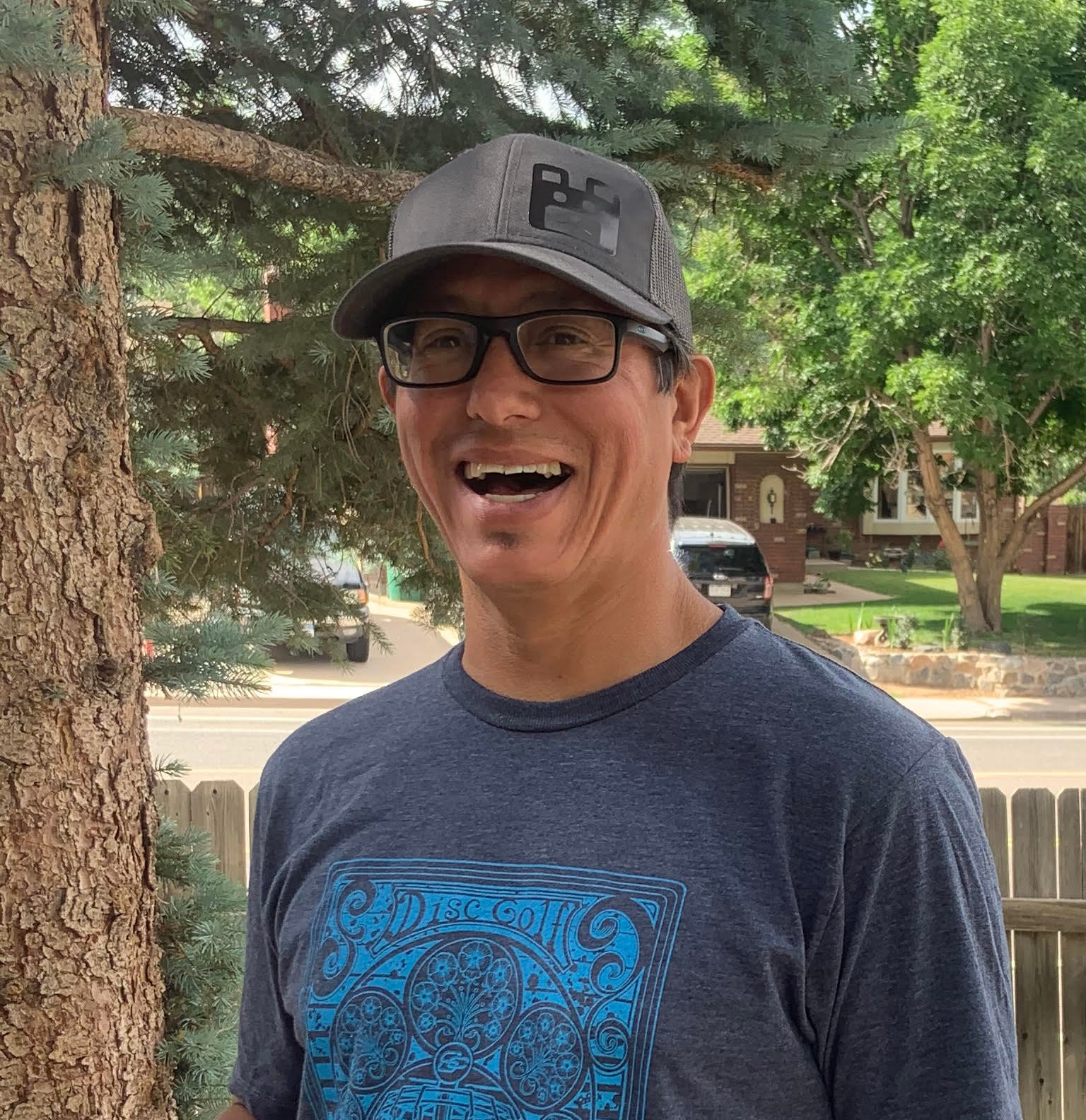 Smiling man wearing glasses and a cap outdoors, trees and a fence in background.