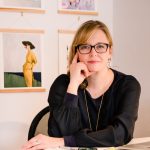 Woman with glasses sitting at a desk, resting her chin on her hand, with framed photos on the wall behind her.