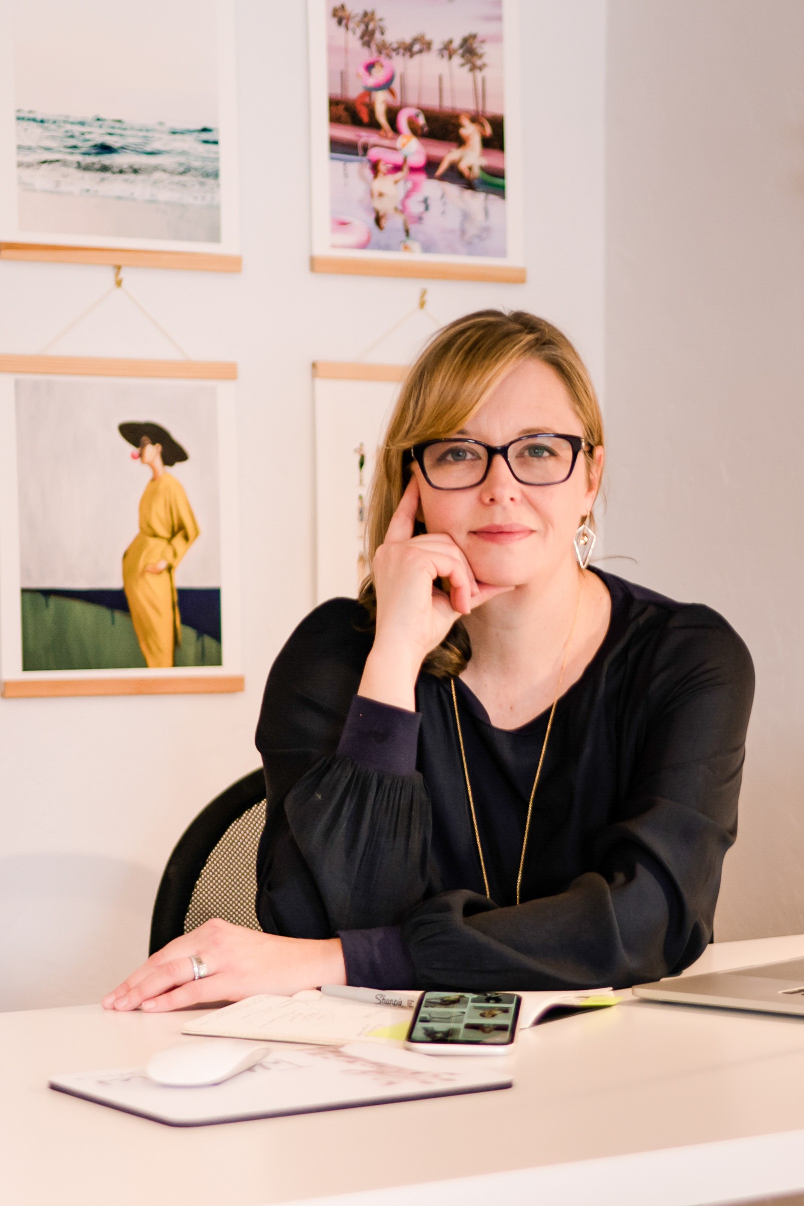 Woman with glasses sitting at a desk, resting her chin on her hand, with framed photos on the wall behind her.