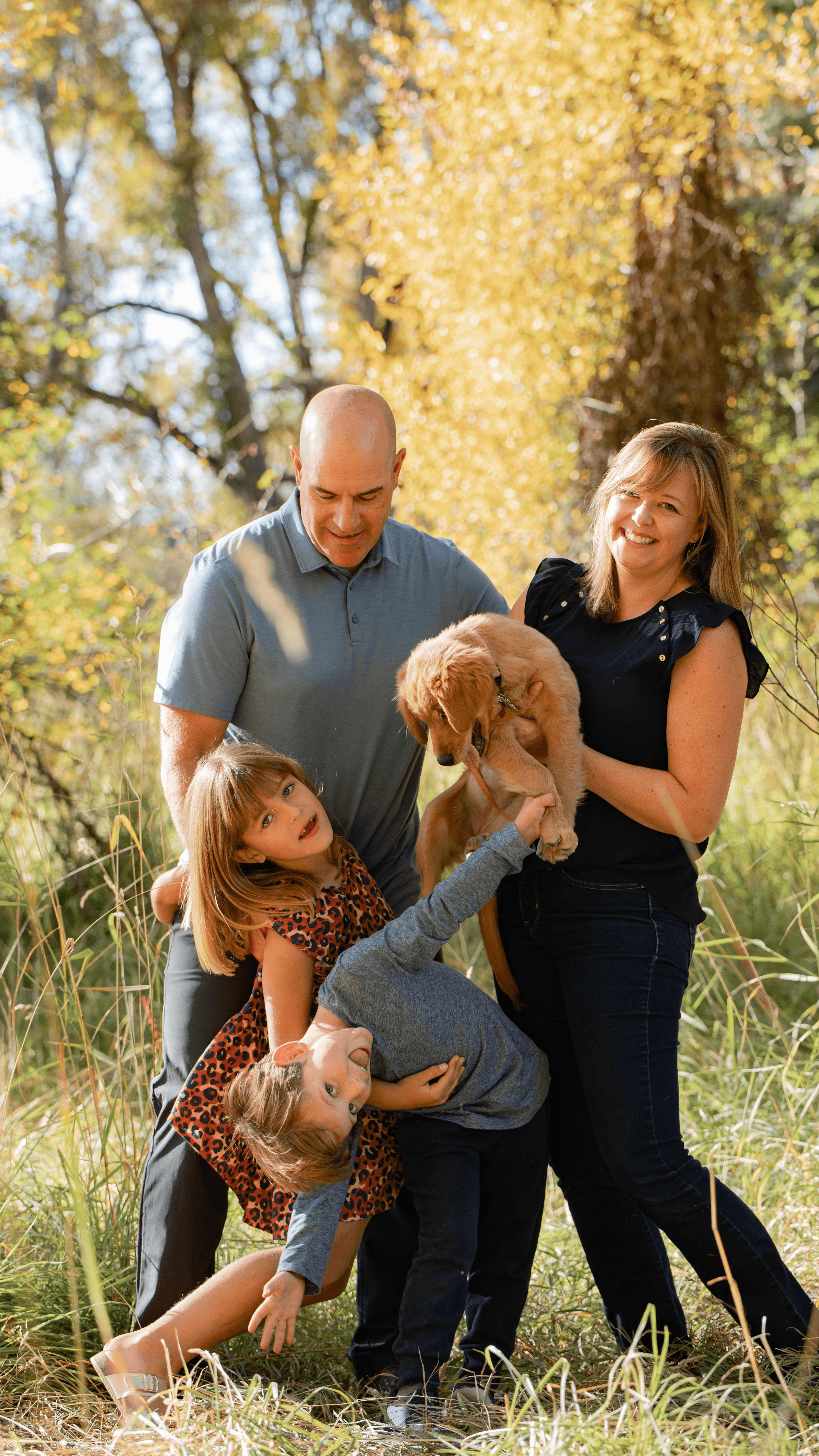 Family of four outdoors with a dog, smiling and playing in a grassy area with trees and autumn leaves.