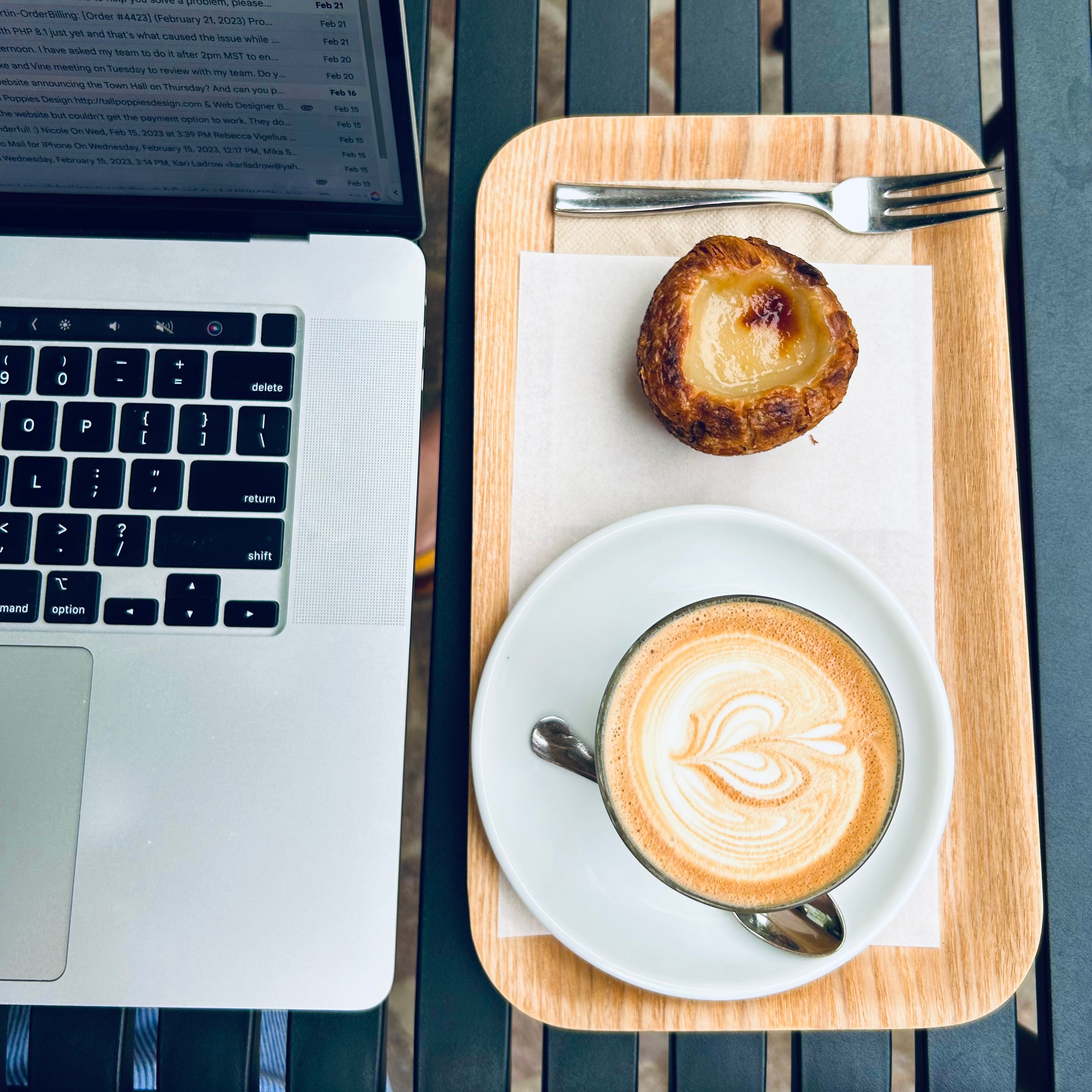 Laptop, coffee, and pastry on a wooden tray with a fork and spoon, on a dark table.