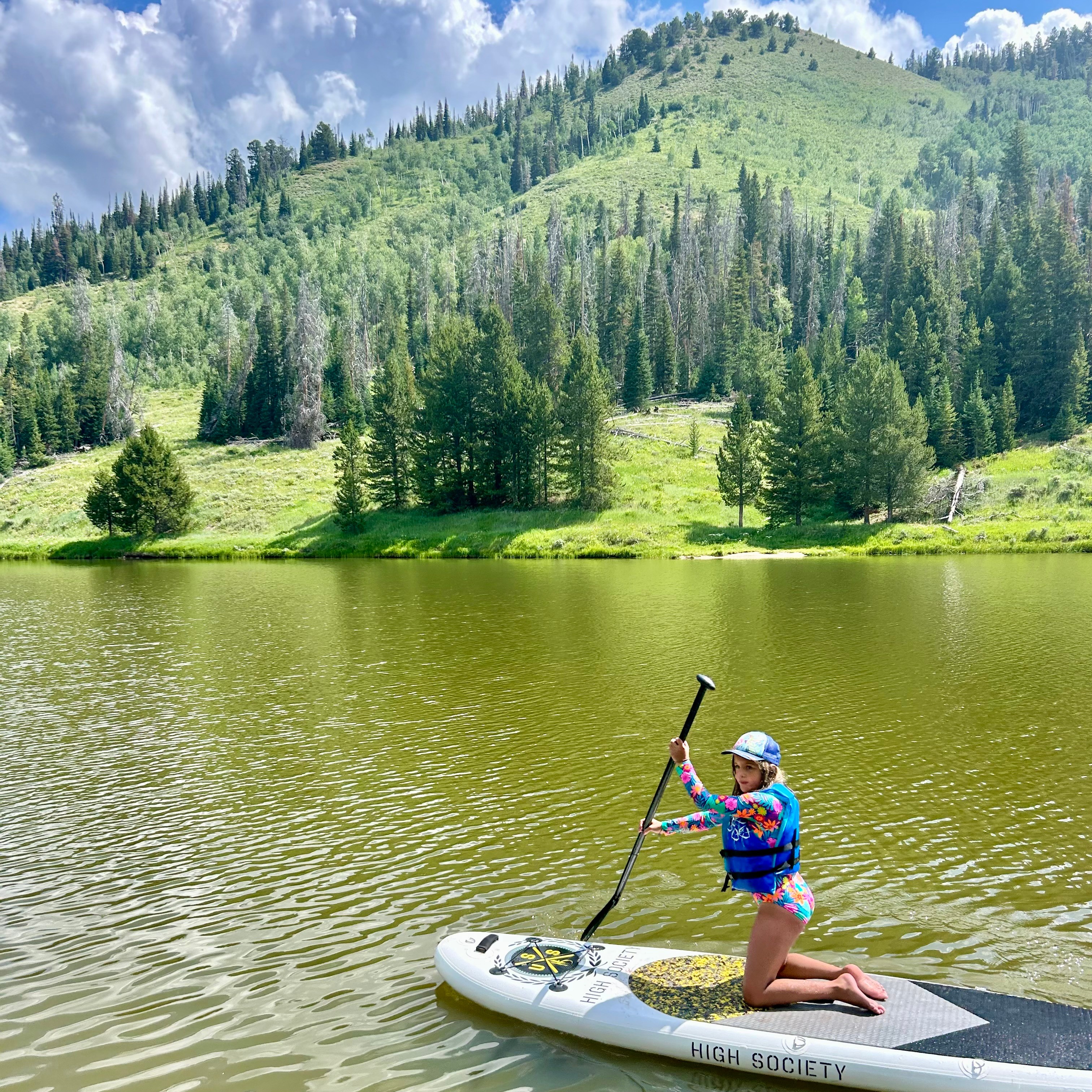 Person kneeling on paddleboard holding paddle near a lake with green hills and trees in the background.