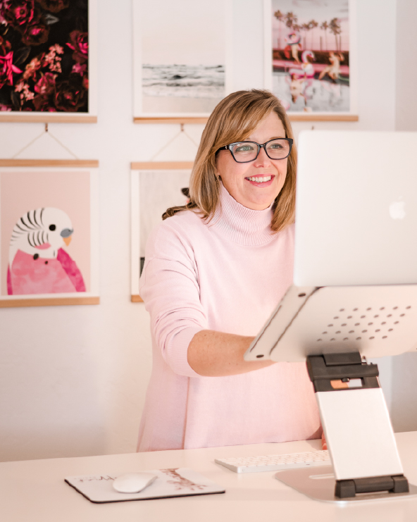 Woman with glasses smiling at a computer in an art gallery, pink sweater, light background, framed artwork on wall