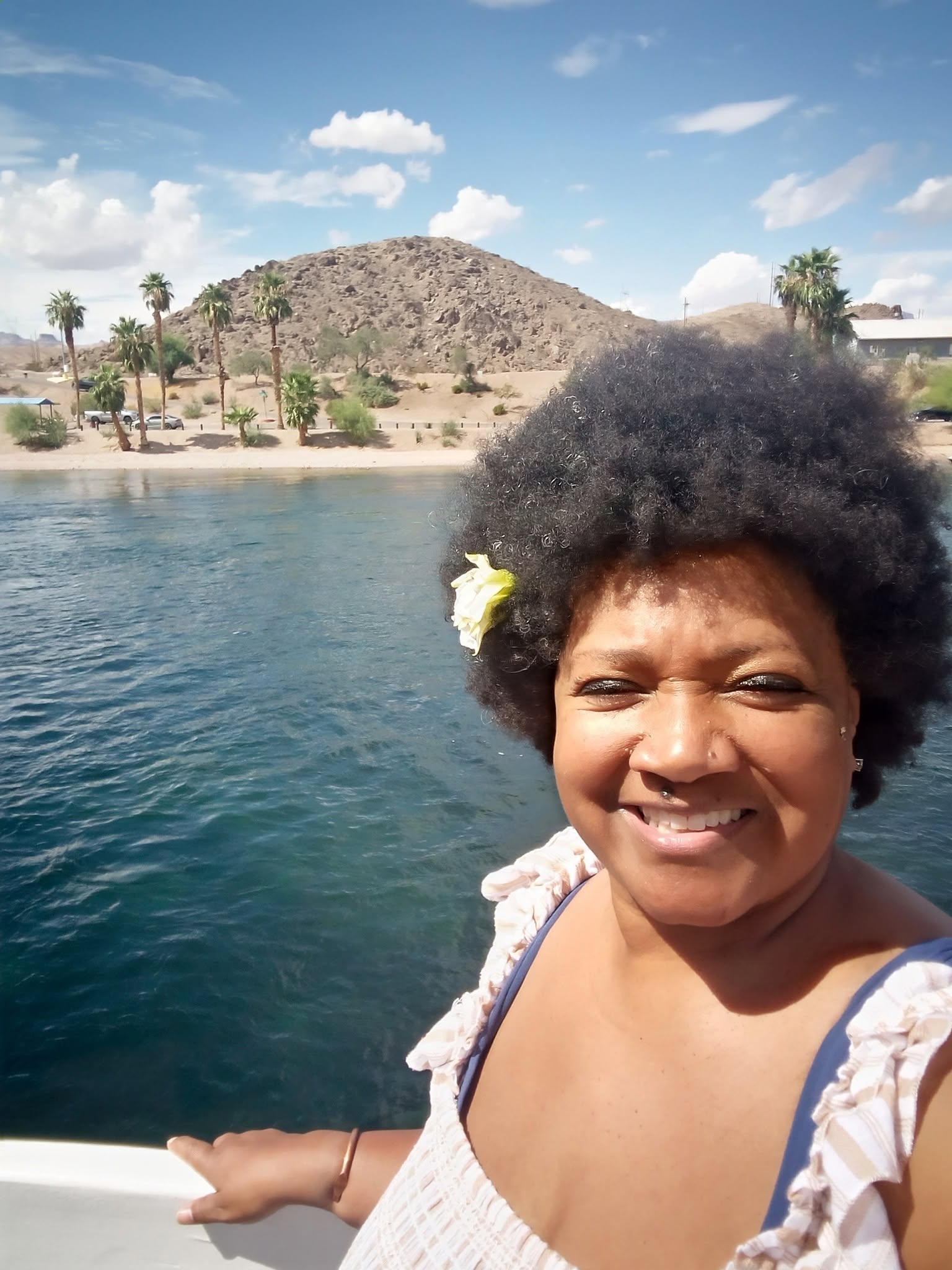 Smiling woman with curly hair and a flower in her hair, standing near water with a hill and trees in the background.