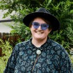 Person wearing a black hat, blue sunglasses, and a patterned shirt outdoors with greenery in the background.