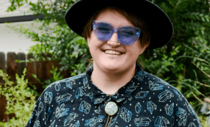 Person wearing a black hat, blue sunglasses, and a patterned shirt outdoors with greenery in the background.