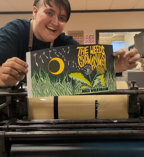Person smiling holding a colorful book titled 'The Weeds Grow Wild' in a room with a window and equipment.