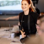 Smiling woman with red hair holding a camera in a meeting room, with a large screen in the background.