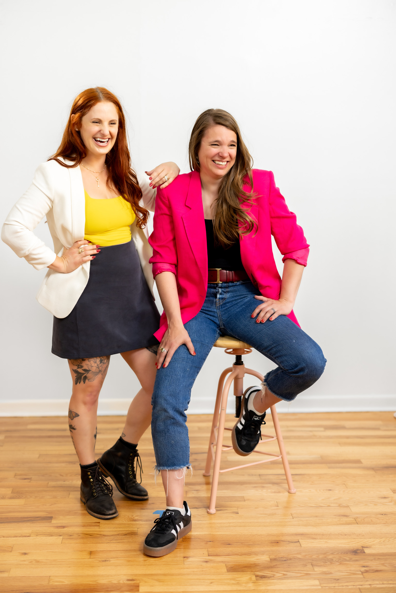 Two women smiling, one standing and one sitting on a stool, against a plain white background.