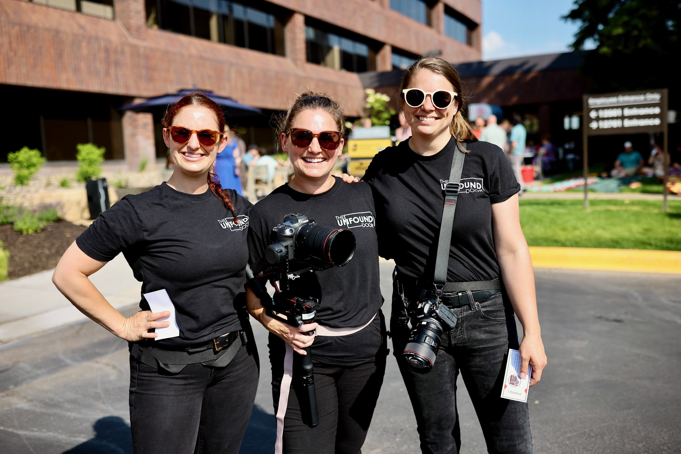 Three women standing outdoors, smiling, wearing black shirts, sunglasses, and carrying cameras, with a building and trees in background.