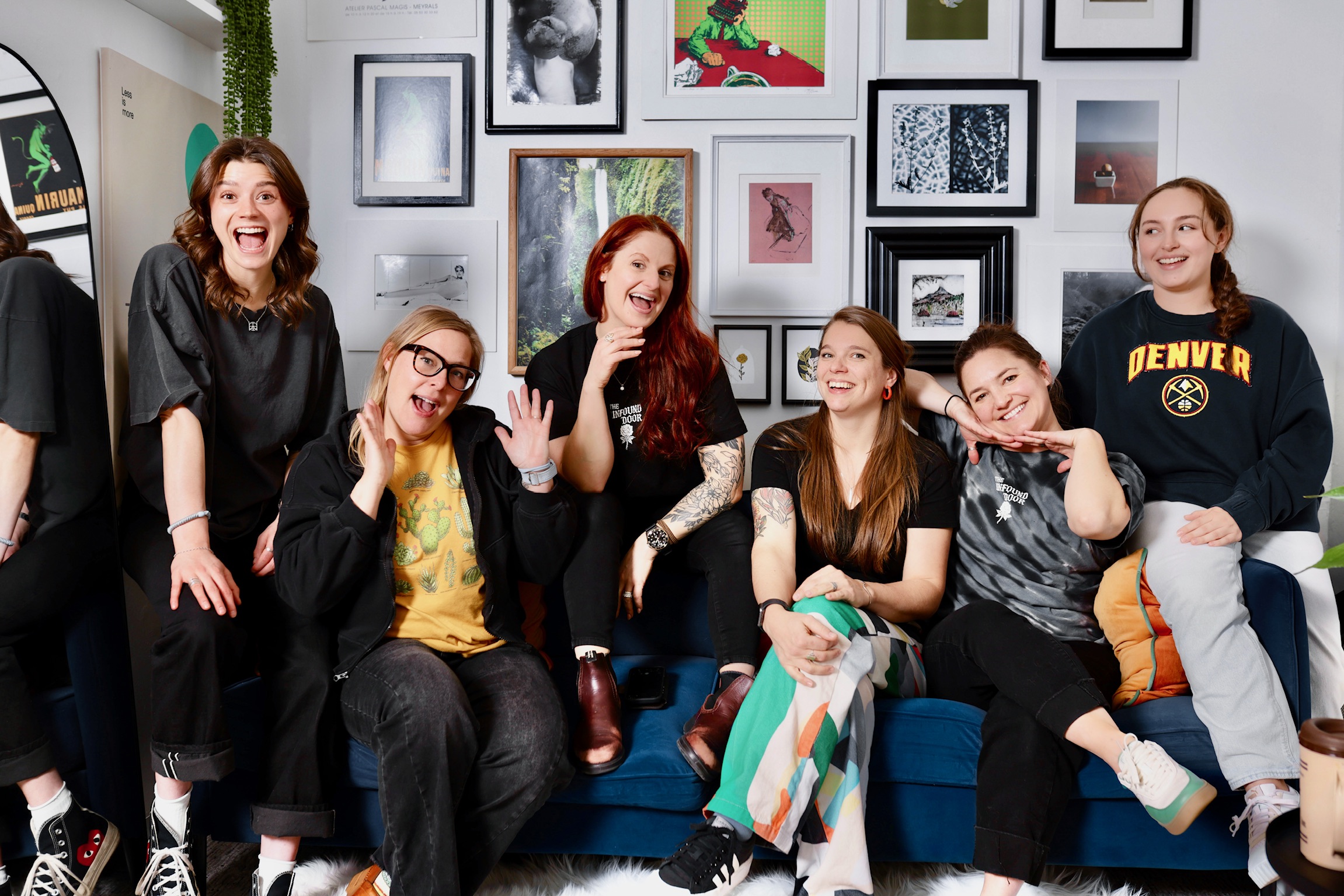 Group of six women sitting and standing on a blue couch, smiling and laughing, with a gallery wall of framed pictures behind them.
