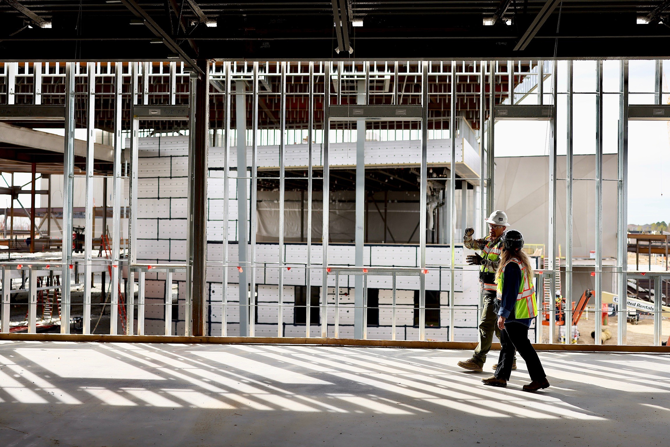 Construction site with metal framework and two workers walking past, partially obscured by shadows and bars.