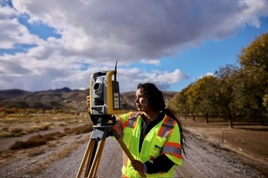 Person in high-visibility vest using surveying equipment outdoors with trees and mountains in background.