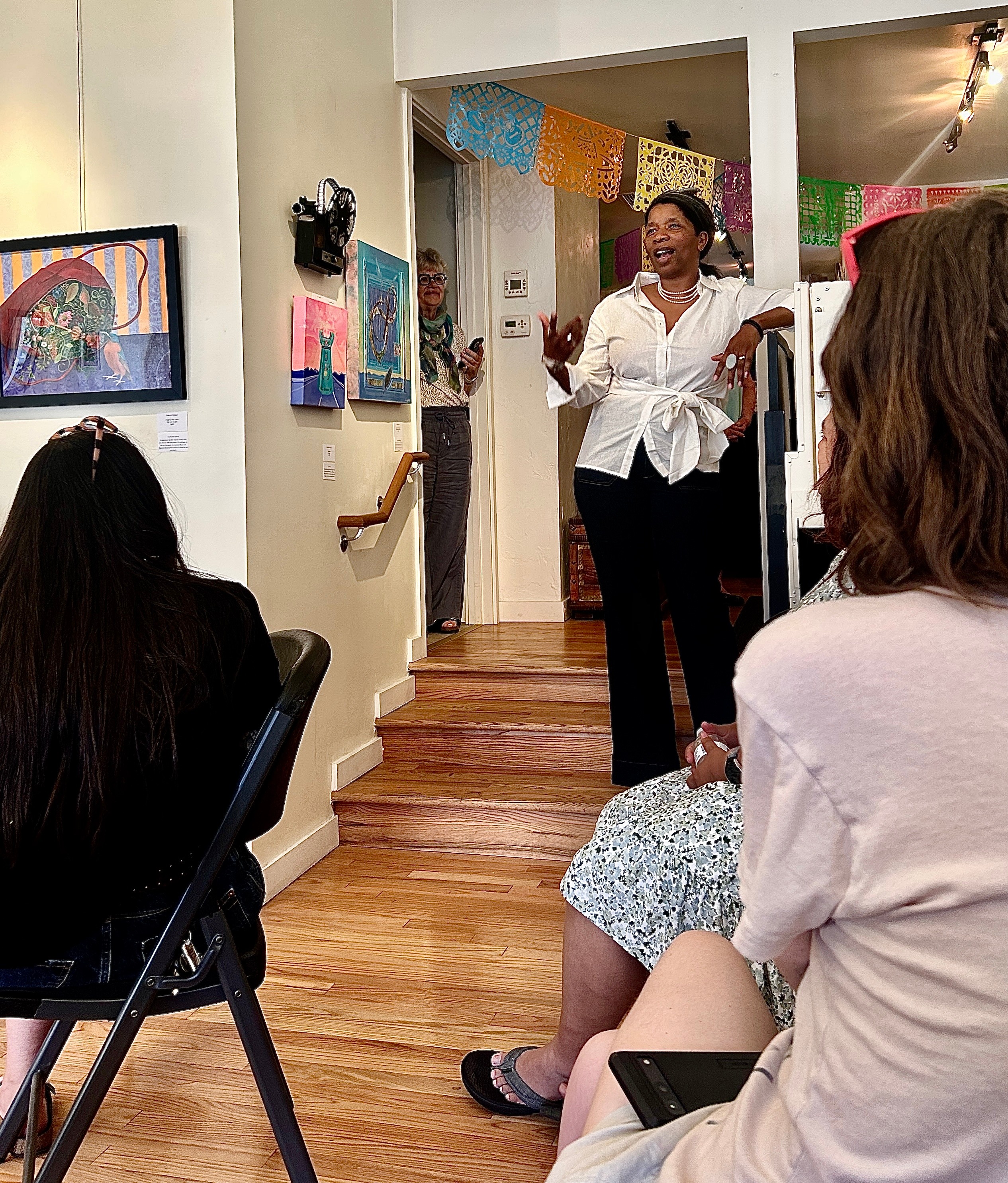 Woman standing and speaking to seated group in a decorated room, colorful banners hanging, artwork on walls, wooden stairs nearby.