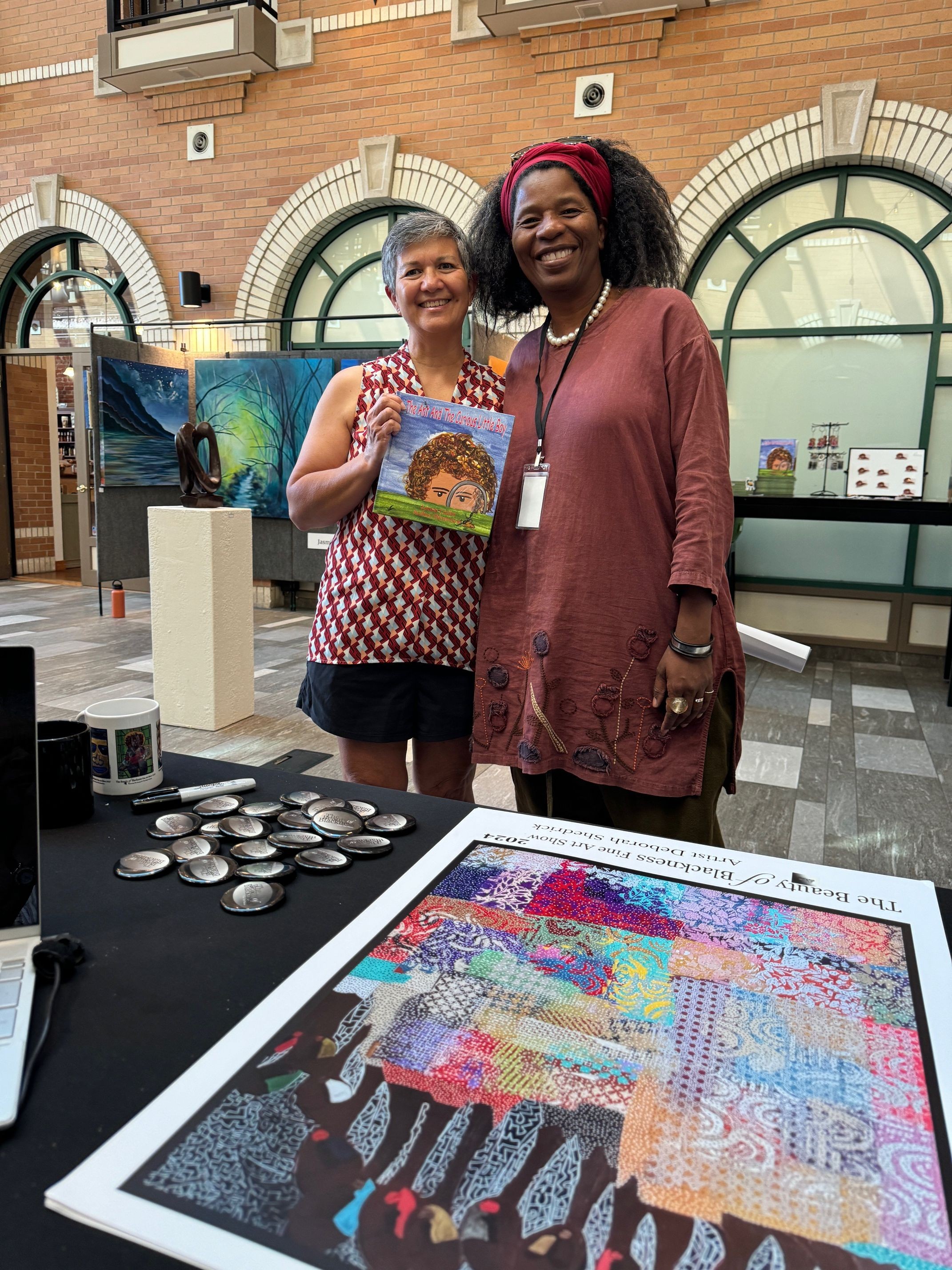 Two women stand behind a table displaying colorful artwork and a digital screen in a gallery setting.