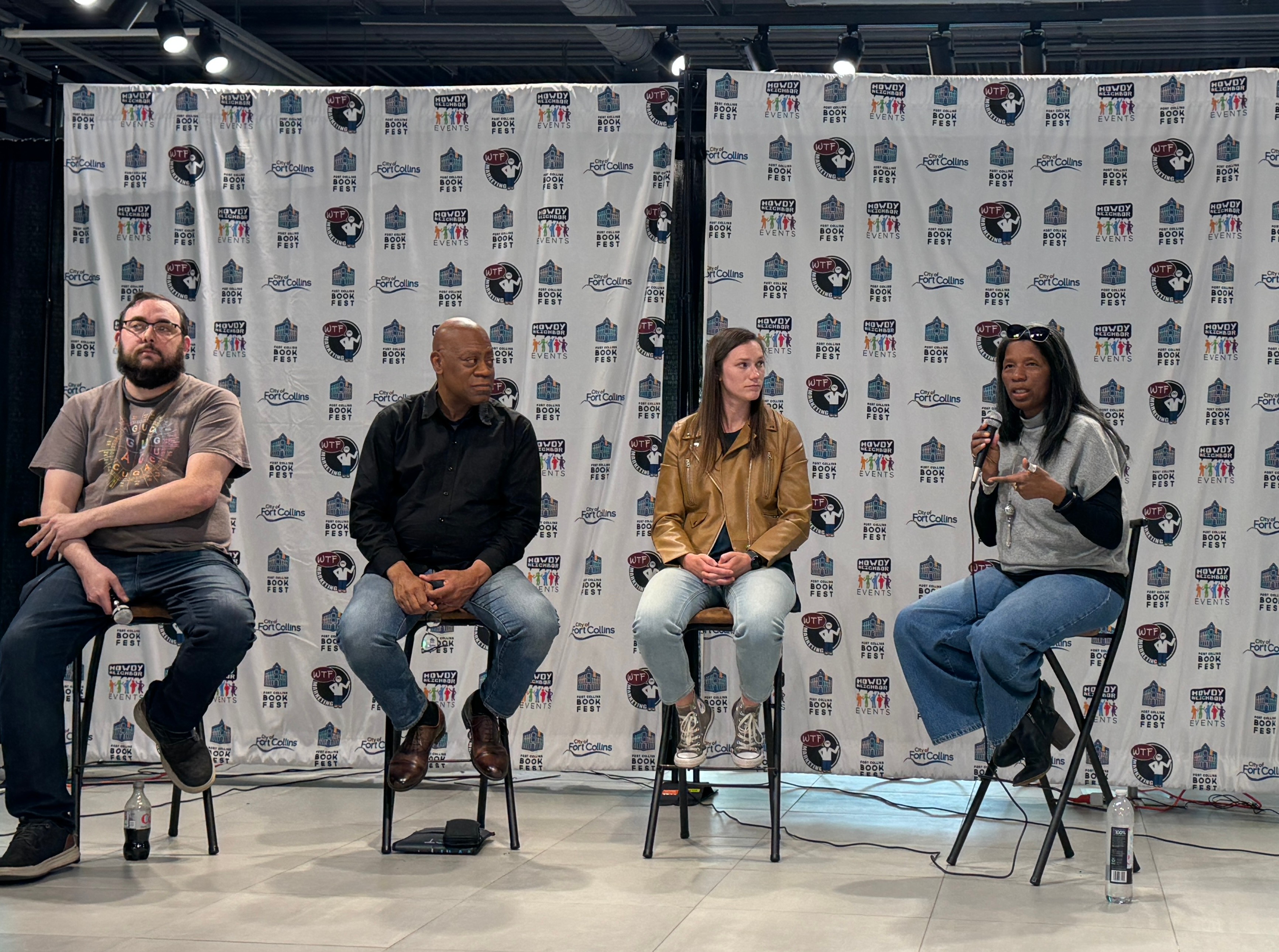 Four people sitting on chairs in front of a backdrop with logos, participating in a panel discussion.