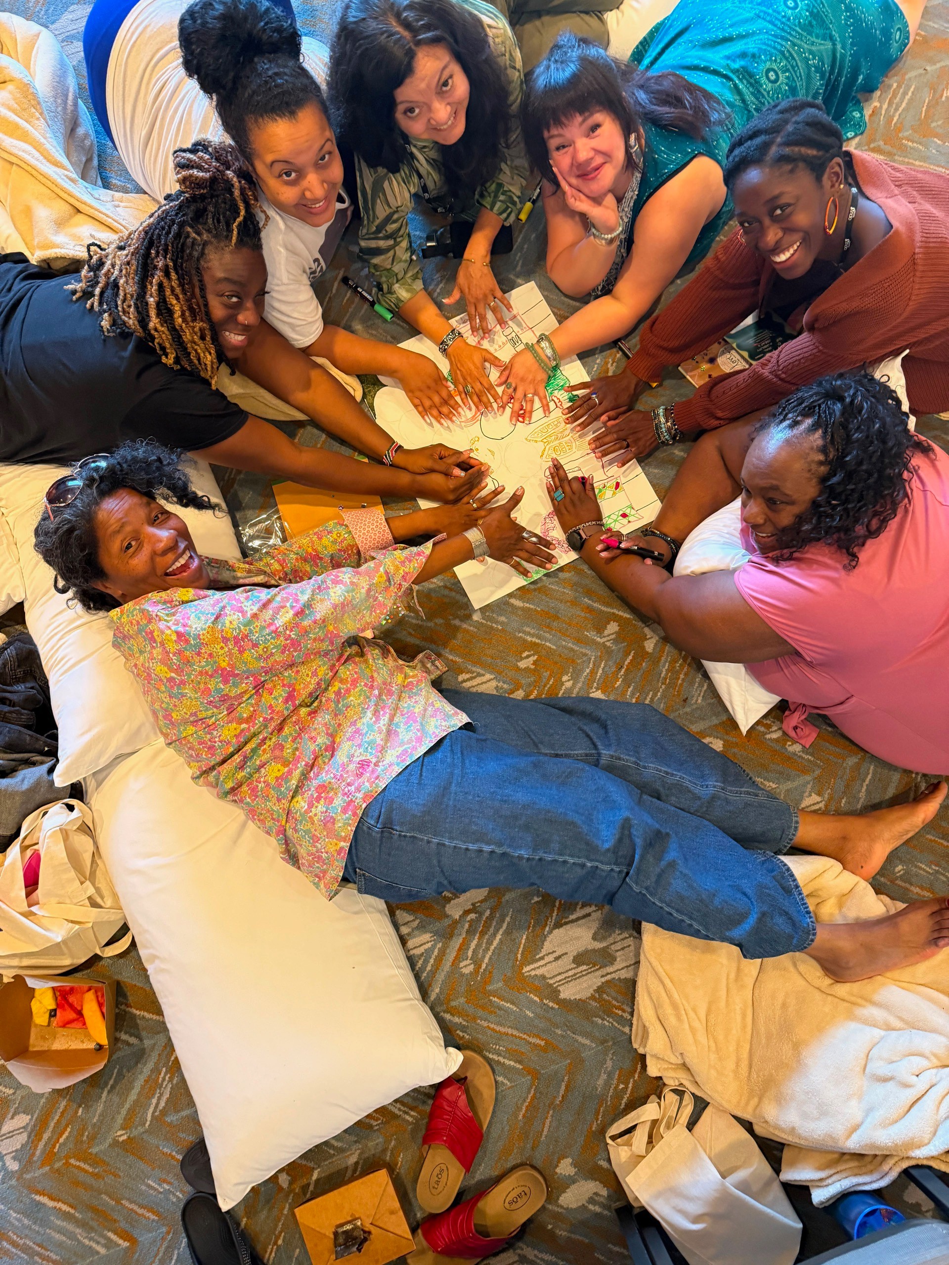 Group of eight diverse women lying on the floor in a circle, holding hands, smiling, and looking up at the camera.