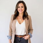 Woman with brown hair smiling, wearing a beige blazer, white top, and dark jeans, standing against a plain background.