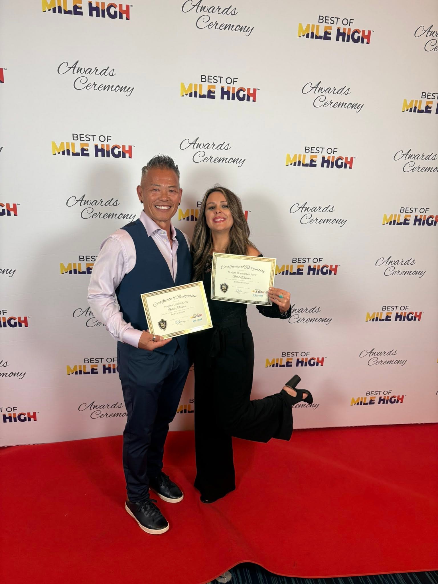 Two people holding certificates on a red carpet, smiling, with a backdrop featuring event logos and text.
