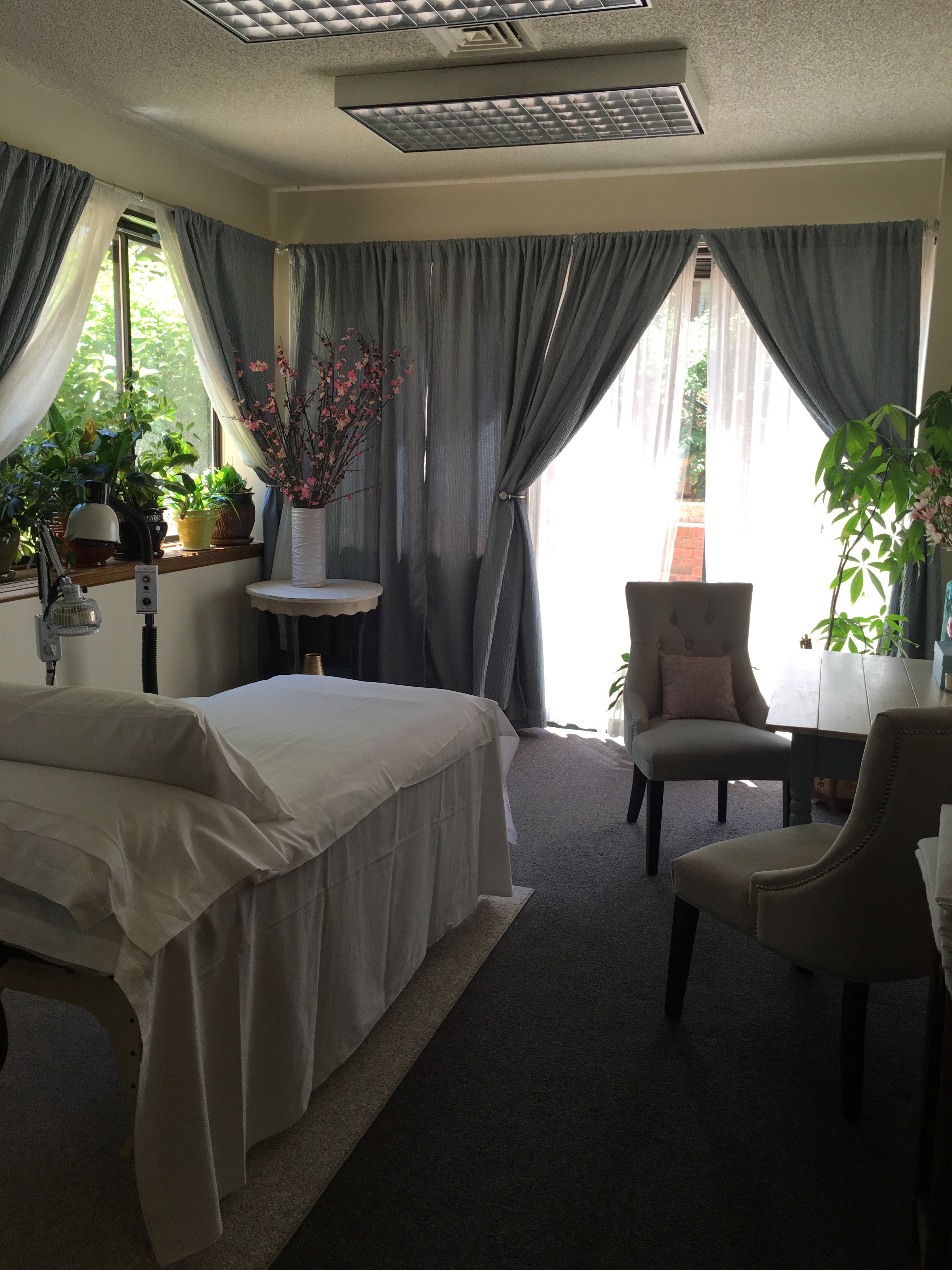 A bedroom with a bed, chairs, and large windows with curtains, sunlight, and plants inside.