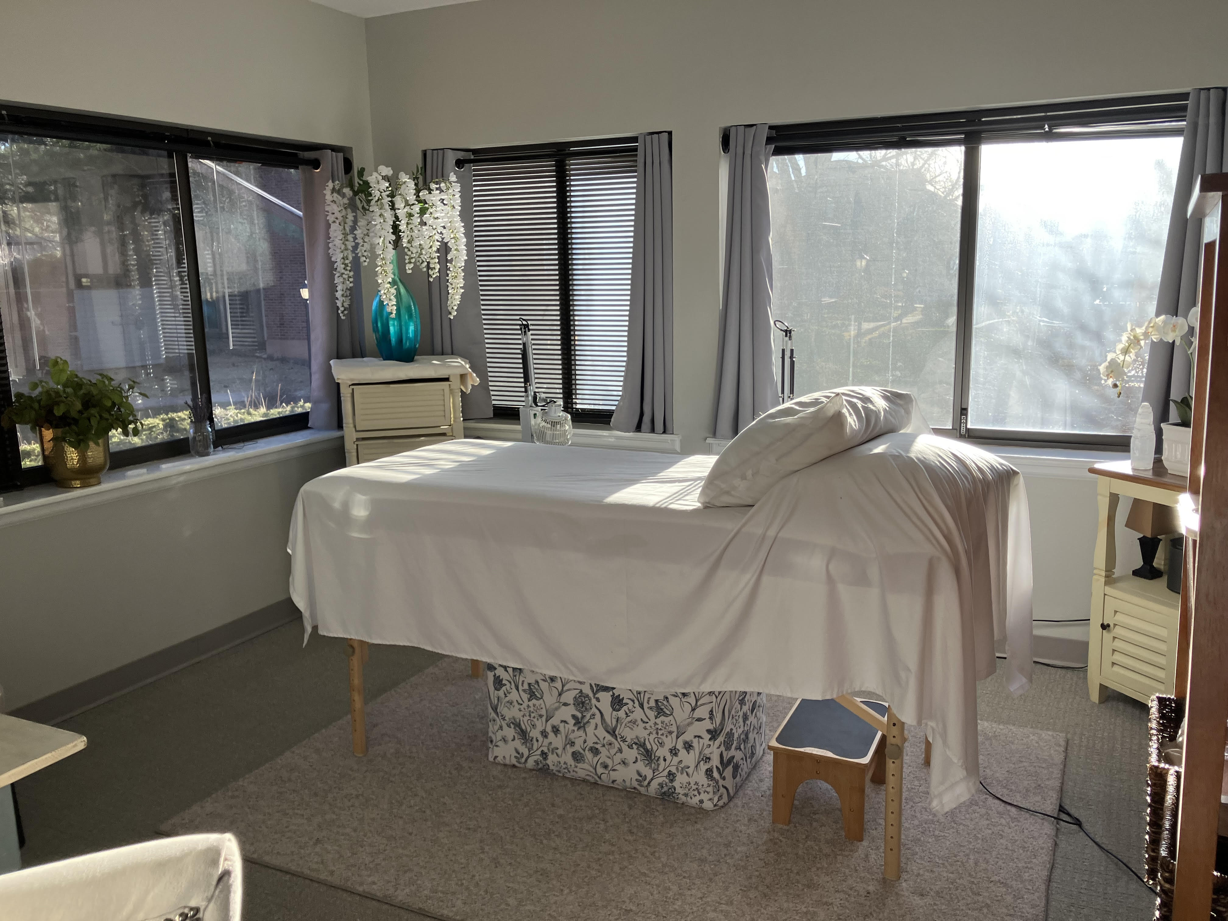 Room with massage table covered in white sheet, windows with blinds and curtains, and decorative plants and furniture.