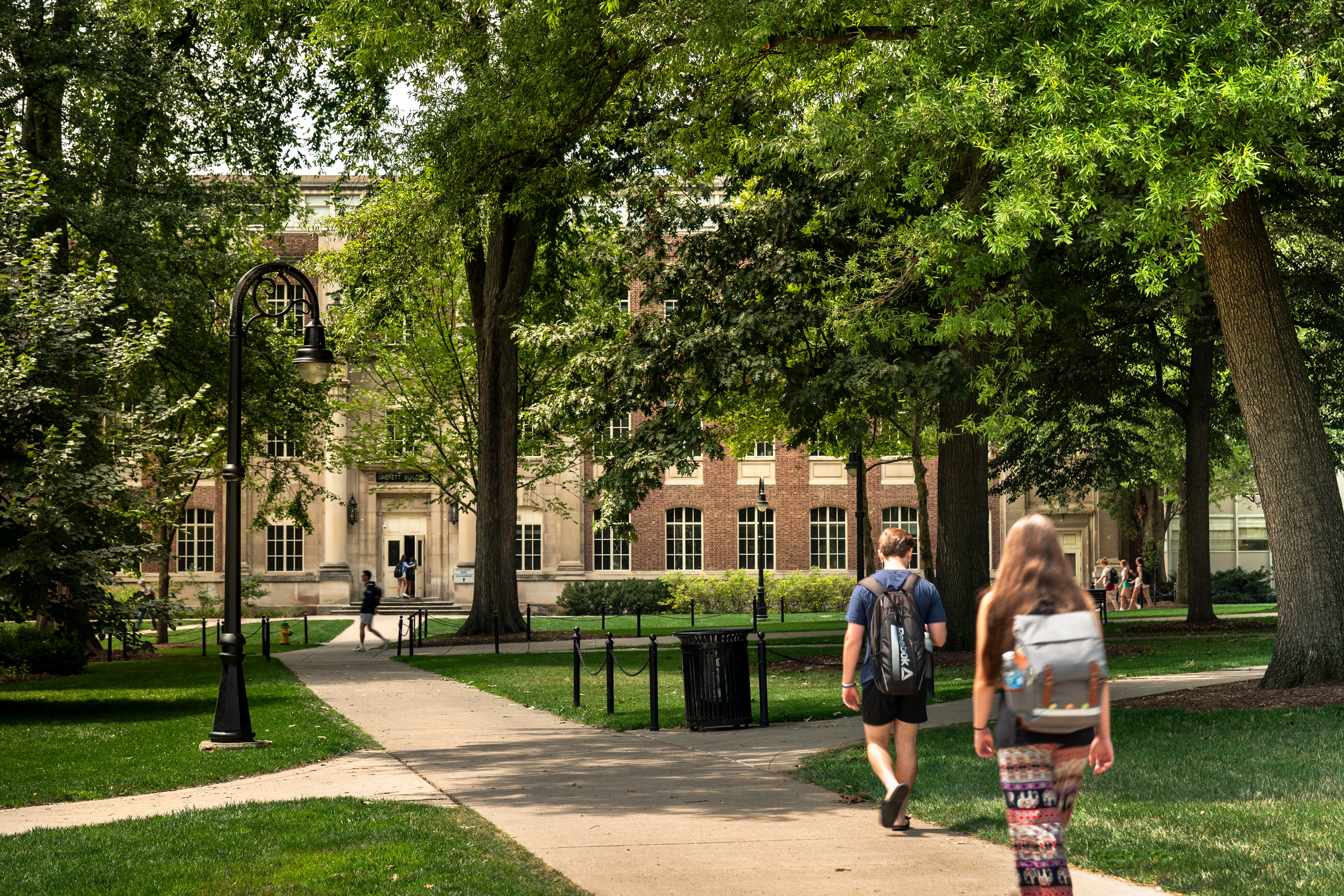 People walking in a park with trees and a building in the background, sunny day, lush green grass.
