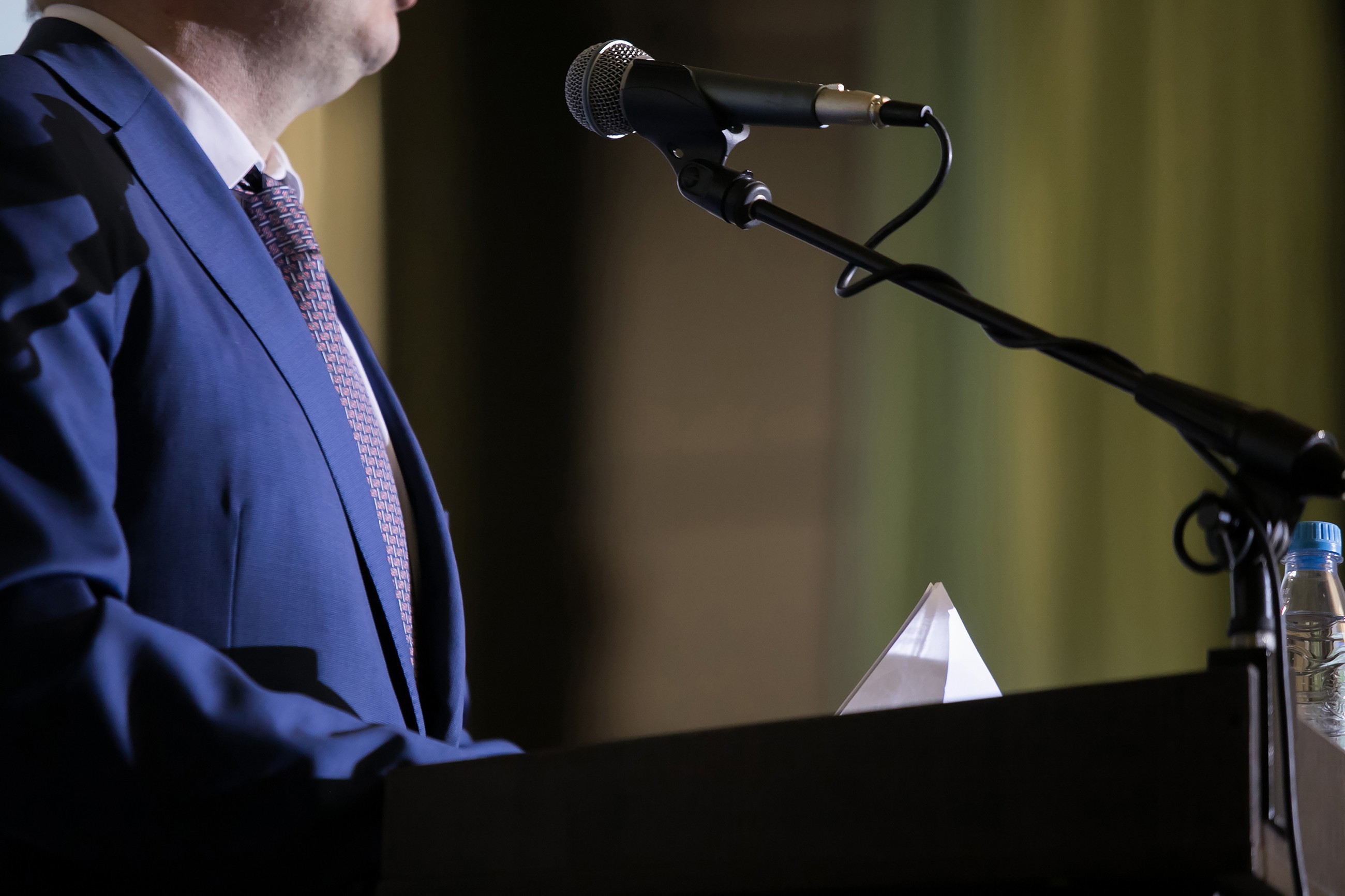 Person in a blue suit speaking at a podium with a microphone, holding papers, with a water bottle nearby.