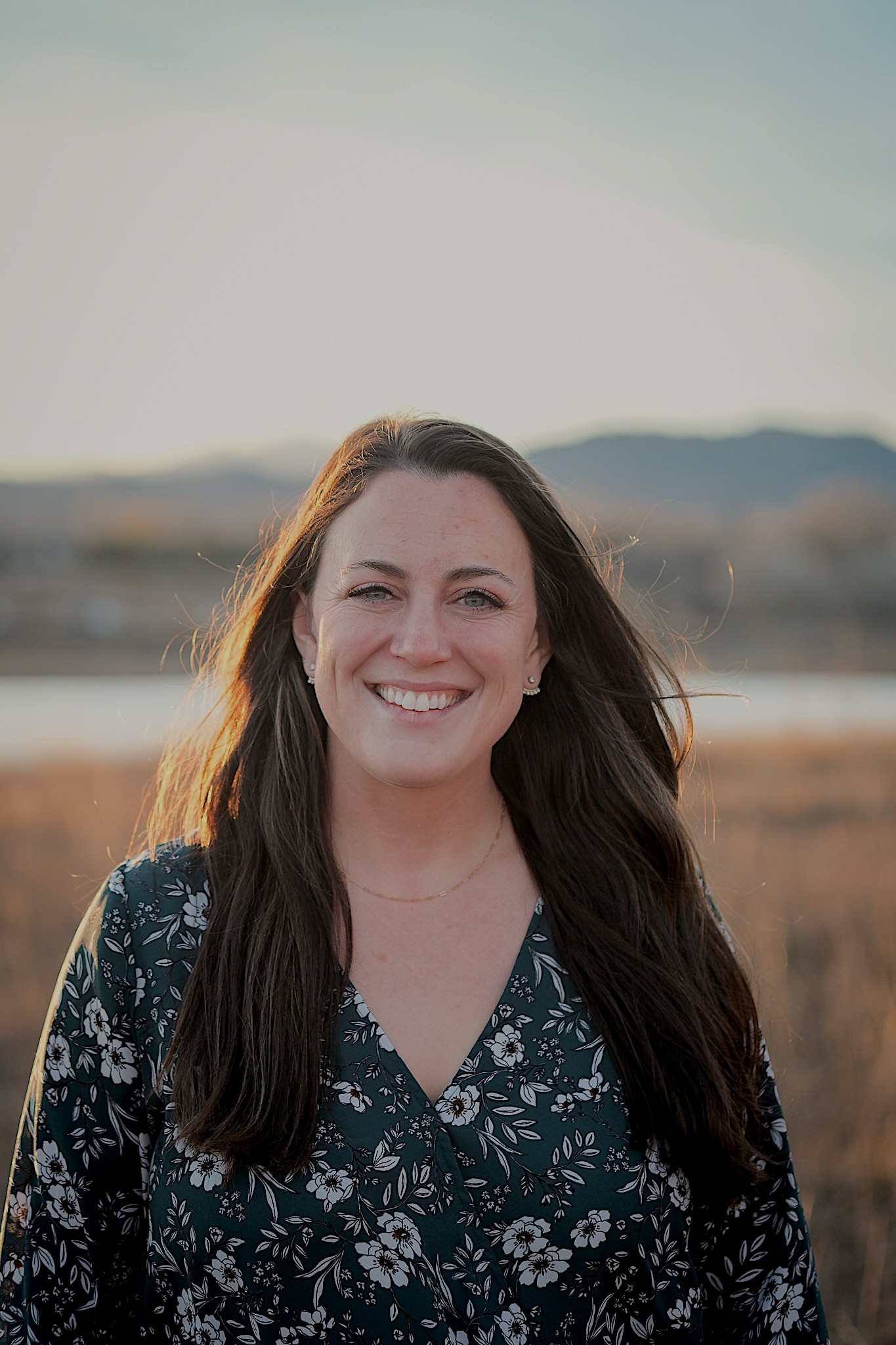 Woman with long dark hair smiling outdoors with a landscape background.