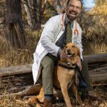 Man with glasses and a beard, wearing a white coat and stethoscope, sitting outdoors with a golden retriever dog, autumn trees in background.