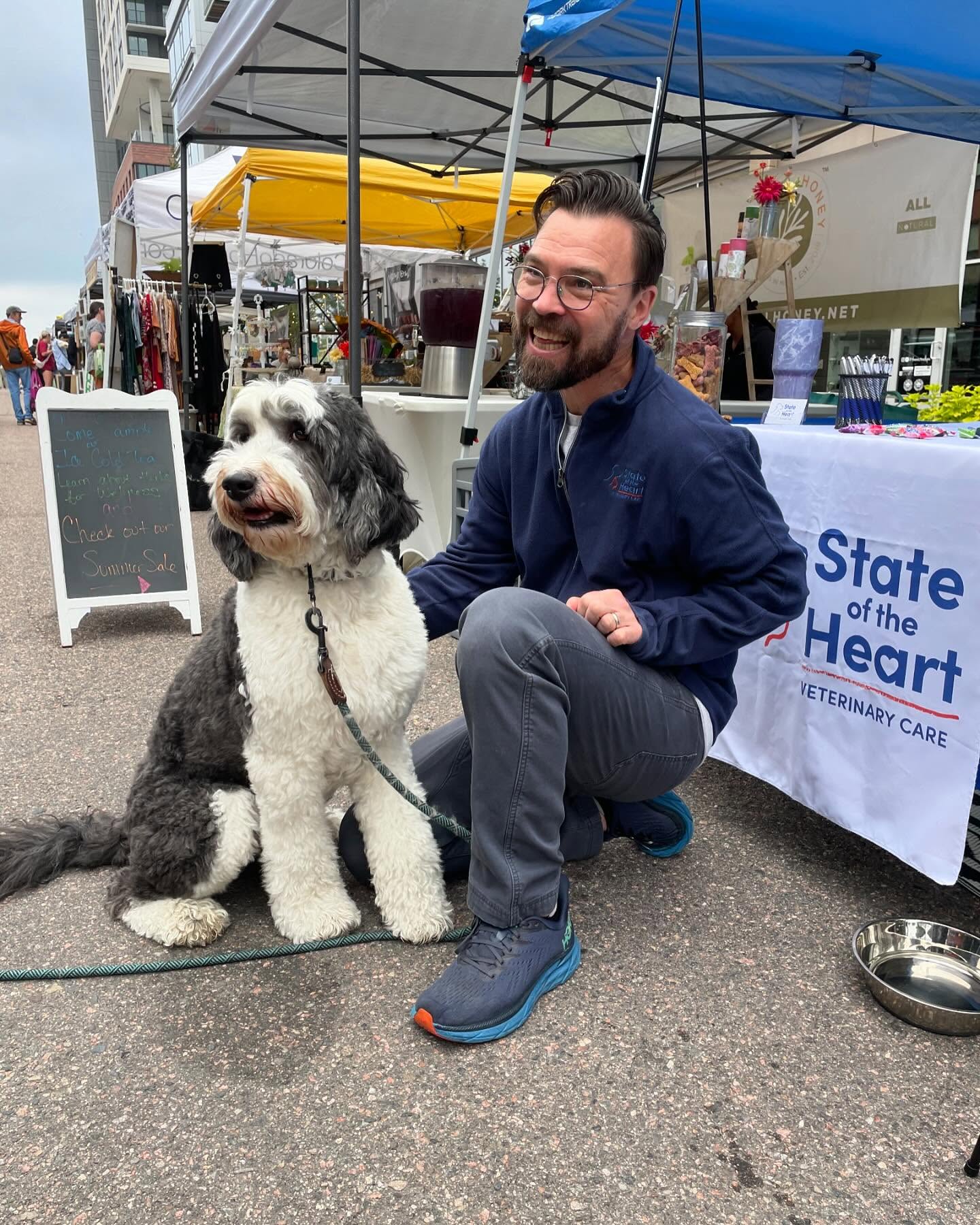 Man kneeling next to a large black and white dog at an outdoor event, with tents and a table in the background.