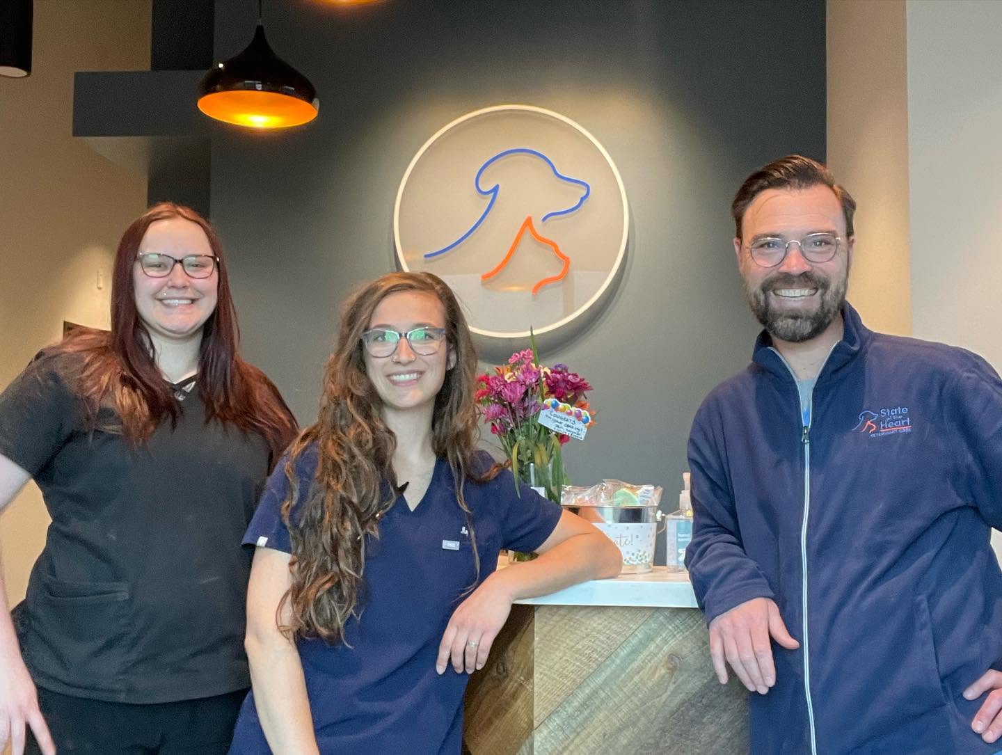 Three people stand in front of a wall with a dog logo, smiling, with a flower arrangement on a counter.