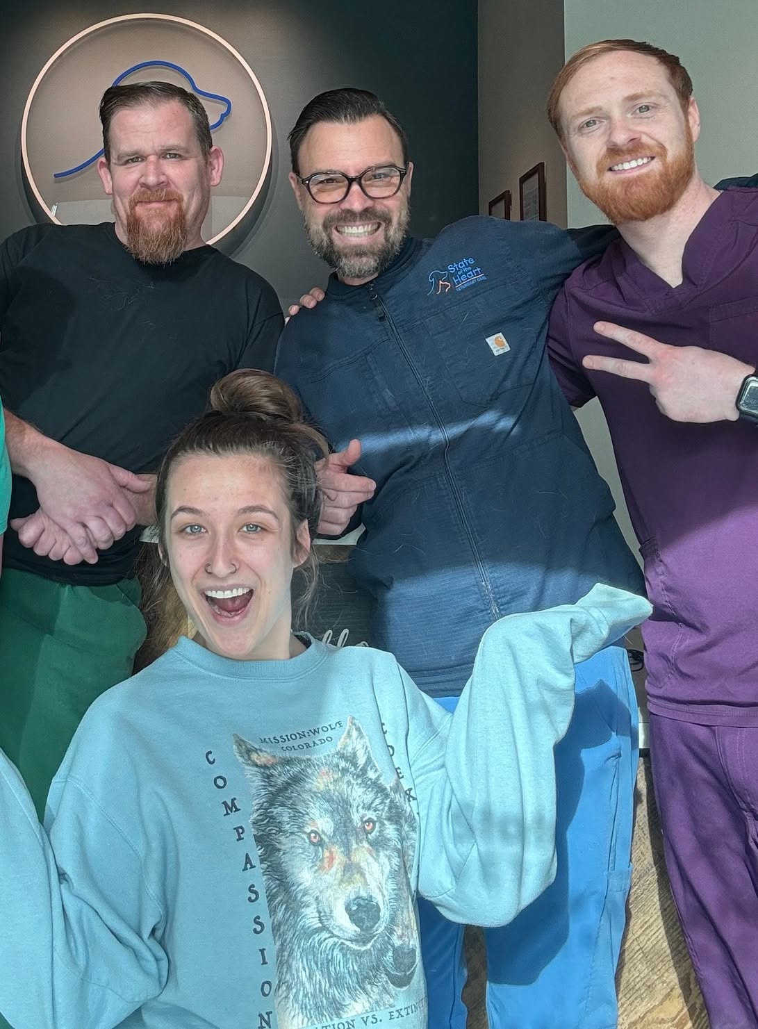 Four people, three men and one woman, smiling indoors, woman holding a light blue sweatshirt, group close together.
