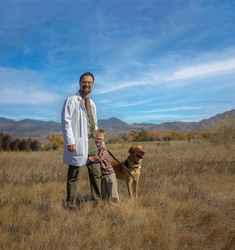 A man in a white coat, a child, and a dog standing in a grassy field with mountains and blue sky in the background.
