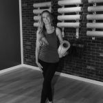 Woman standing in a gym holding a rolled yoga mat, smiling, with wall-mounted exercise equipment behind her.