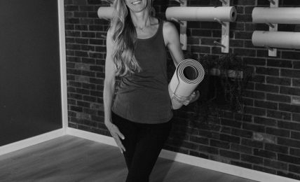 Woman standing in a gym holding a rolled yoga mat, smiling, with wall-mounted exercise equipment behind her.