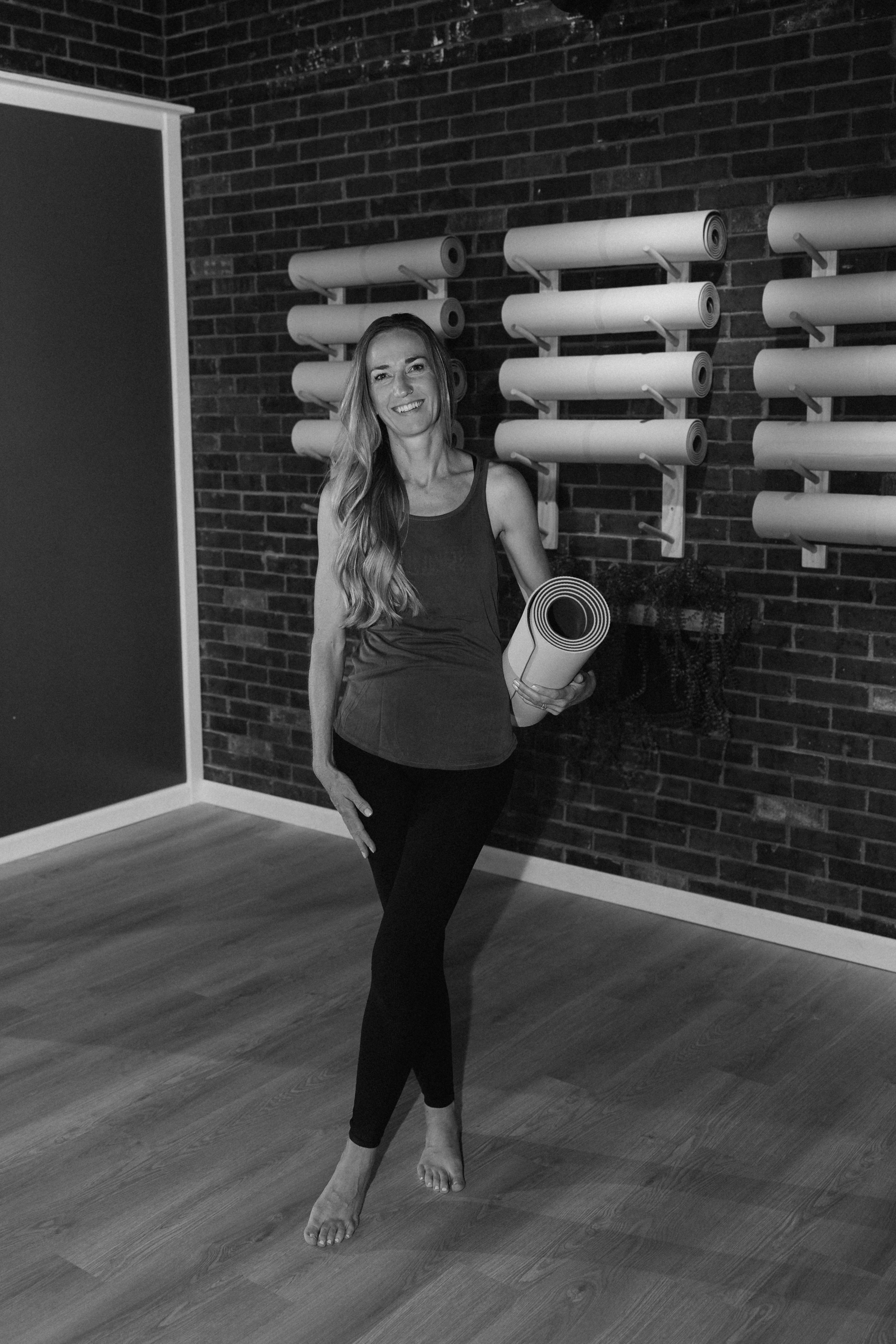 Woman standing in a gym holding a rolled yoga mat, smiling, with wall-mounted exercise equipment behind her.