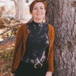 Woman with short hair standing outdoors next to a tree, smiling, wearing a black and white top and brown cardigan.