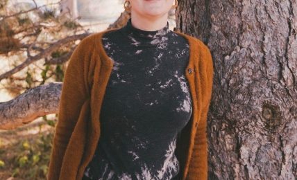 Woman with short hair standing outdoors next to a tree, smiling, wearing a black and white top and brown cardigan.