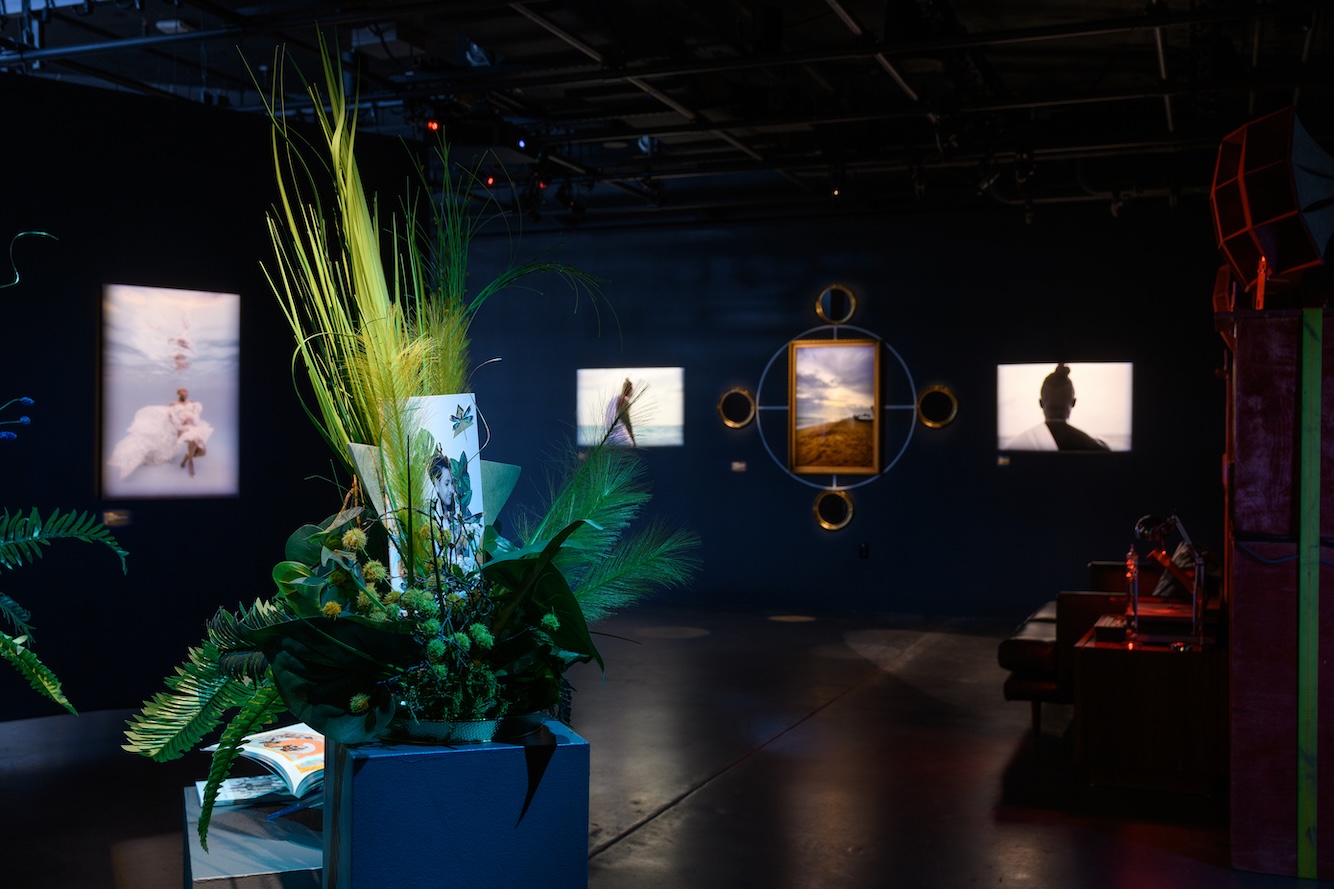 Gallery with illuminated framed photographs on dark walls and a large floral arrangement in the foreground.