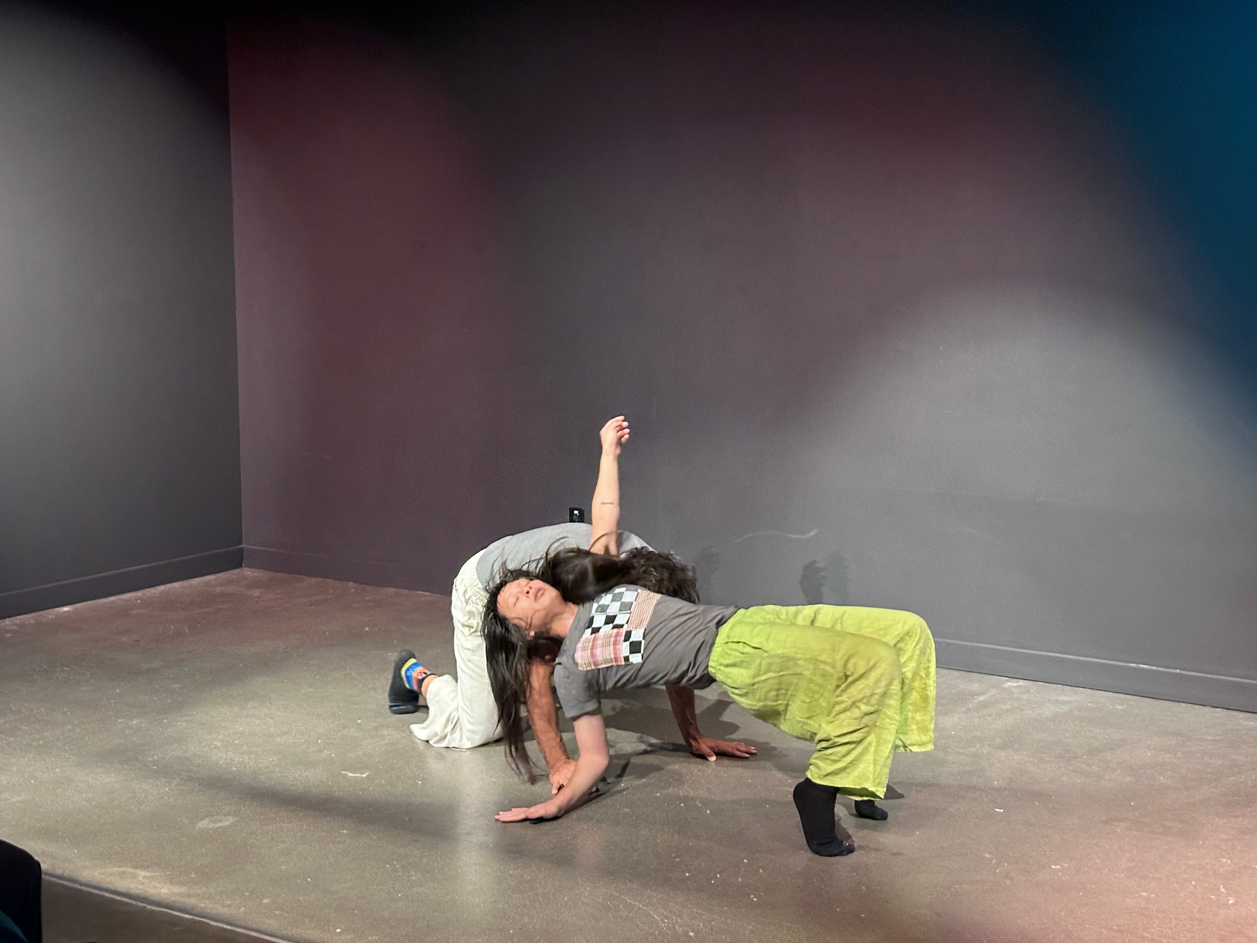 Two children playing on the floor in a room with dark walls, one child on hands and knees, the other reaching upward.