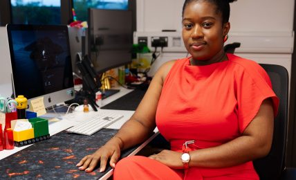Woman in red outfit sitting at a desk with computer and LEGO blocks, in an office with window and TV behind her.