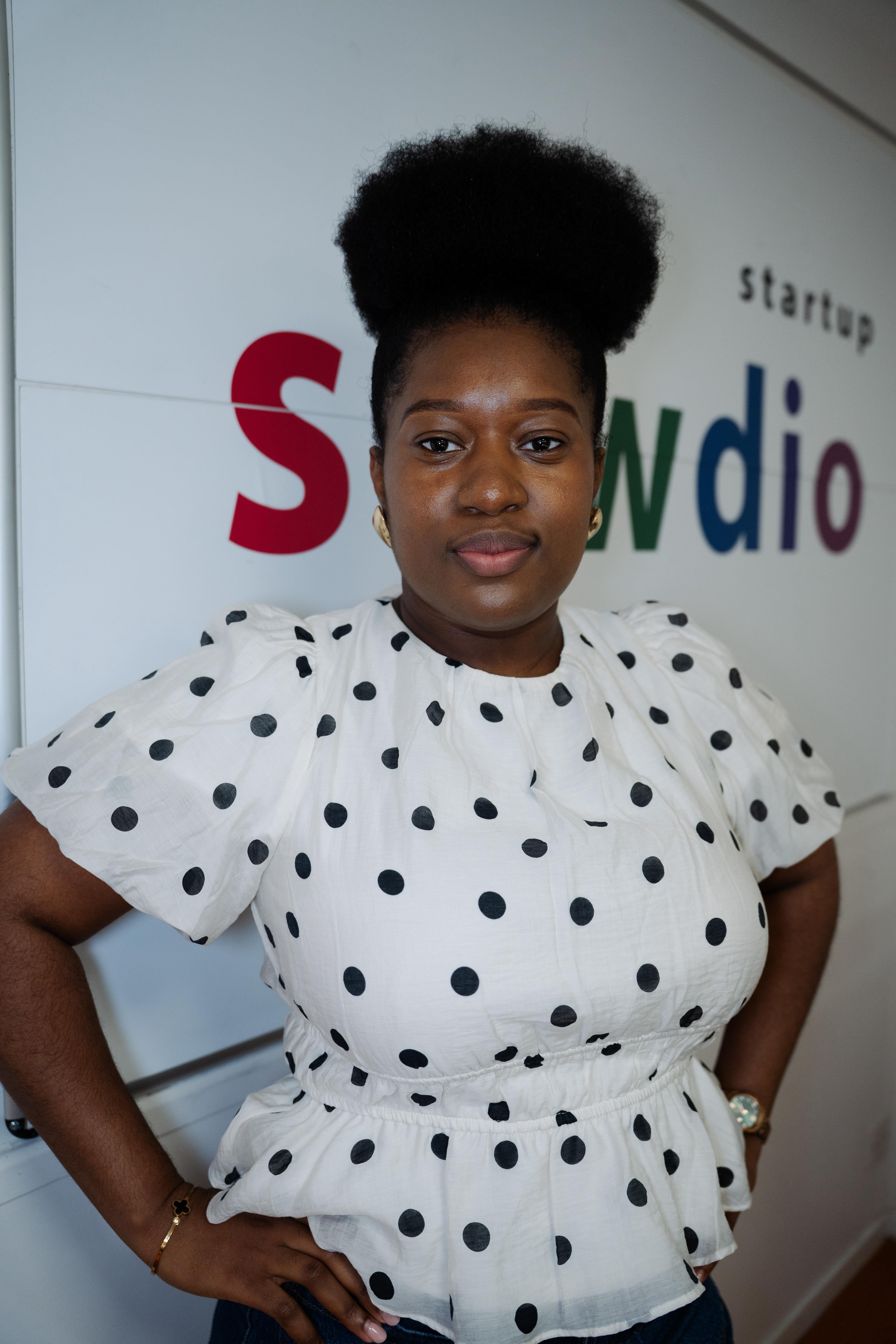 Young woman with natural hair in a polka dot blouse standing indoors near a wall with colorful text.