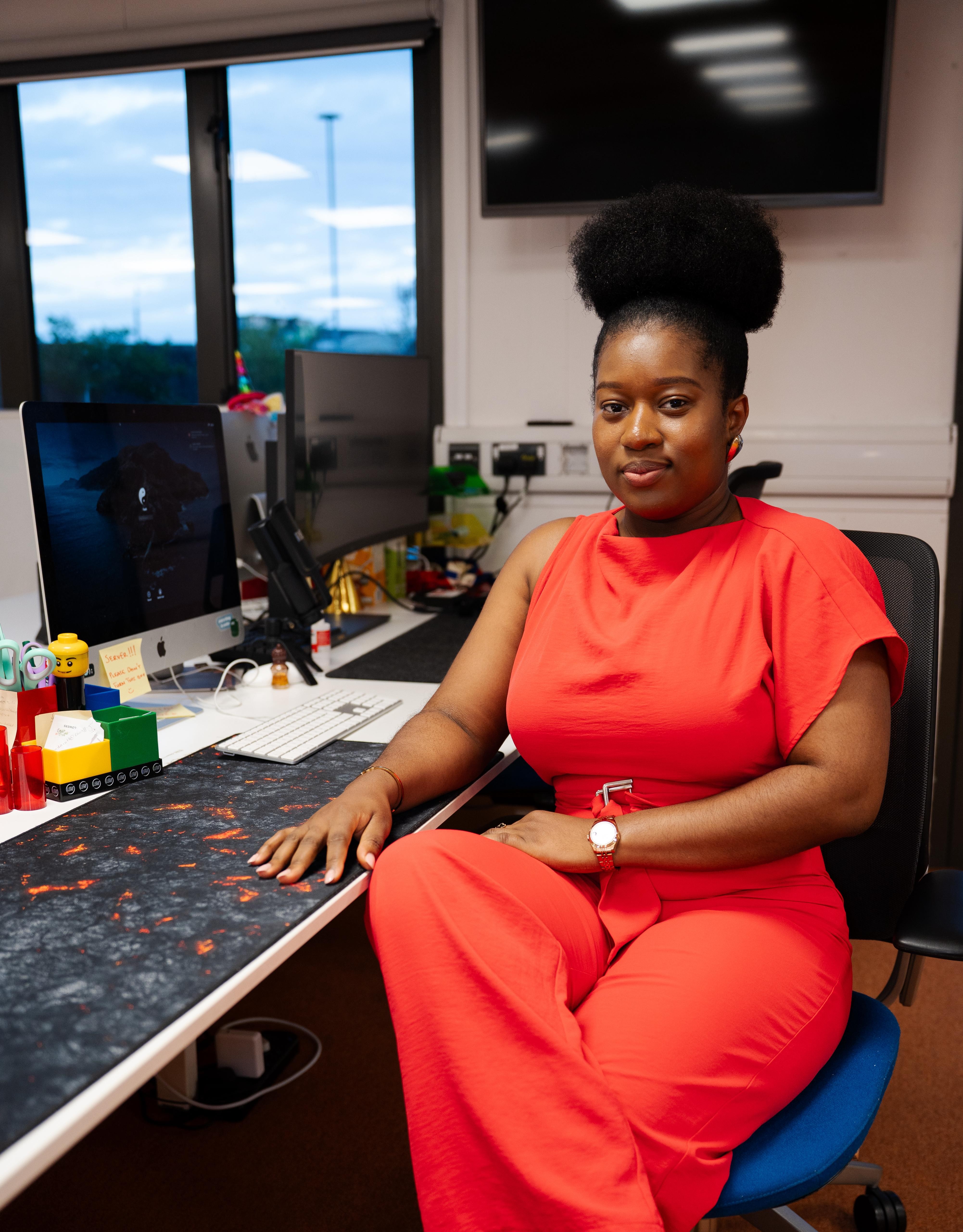 Woman in red outfit sitting at desk with computer and toys, window showing outdoor scene, in office setting.
